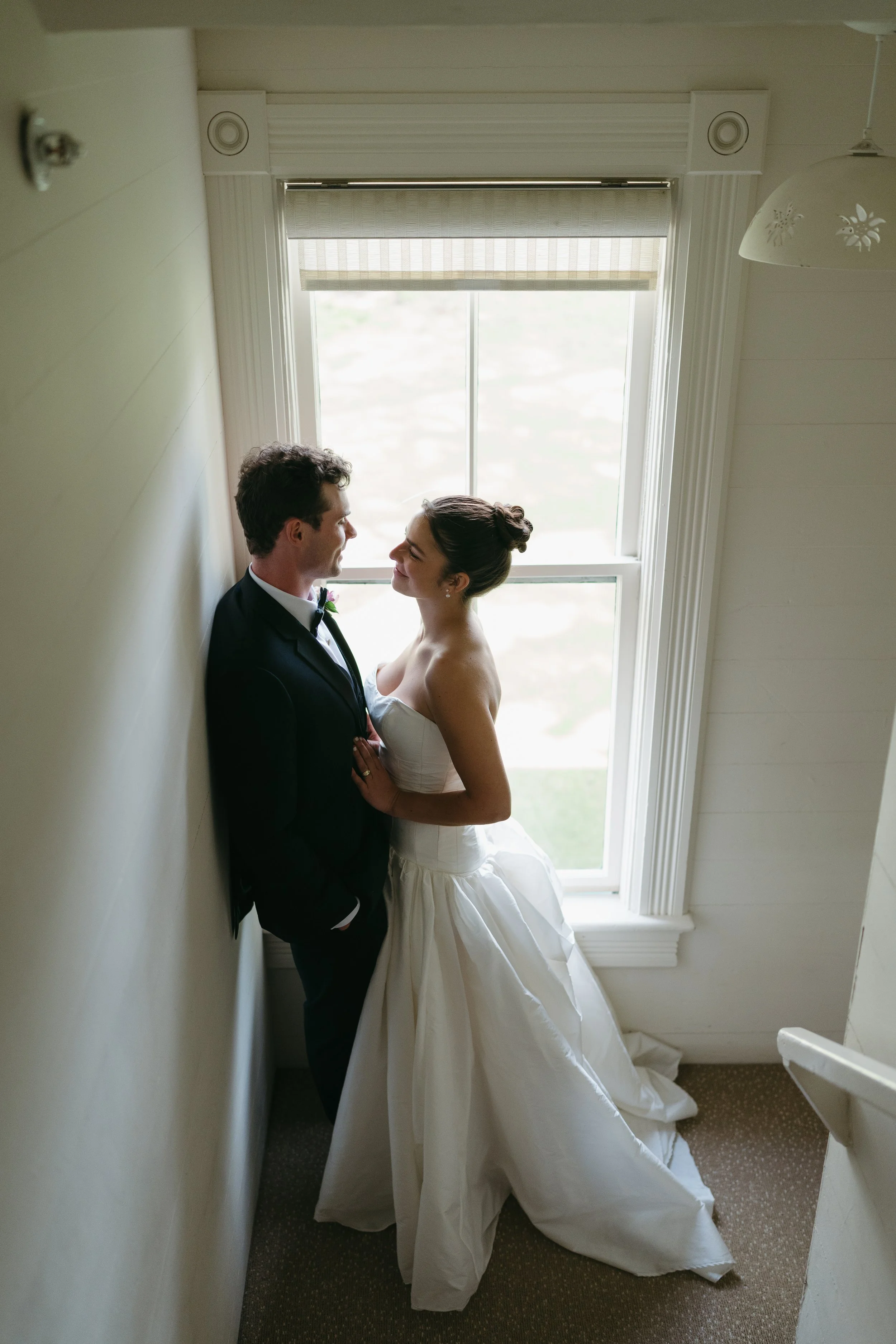 A bride and groom share a quiet moment on a staircase, smiling at each other in front of a softly lit window. The bride wears a strapless white gown, and the groom wears a black tuxedo.