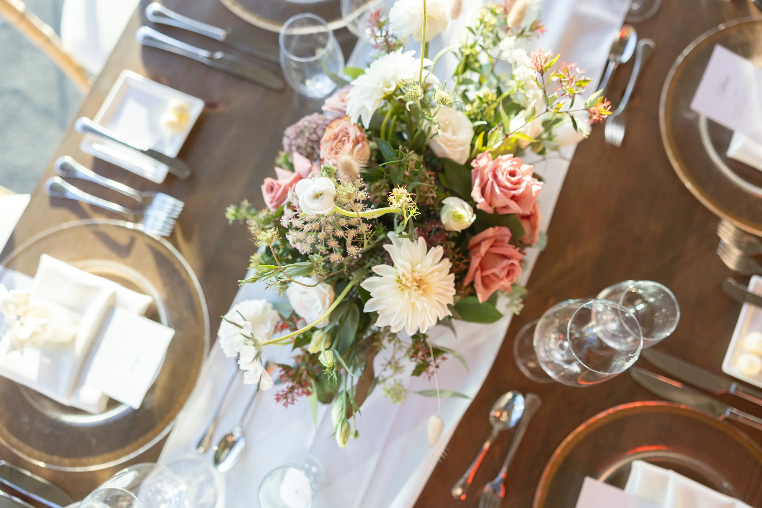 Overhead view of a wedding table with a centerpiece of blush and coral roses, cream dahlias, and greenery, surrounded by gold-rimmed chargers, white napkins, silverware, and glassware.