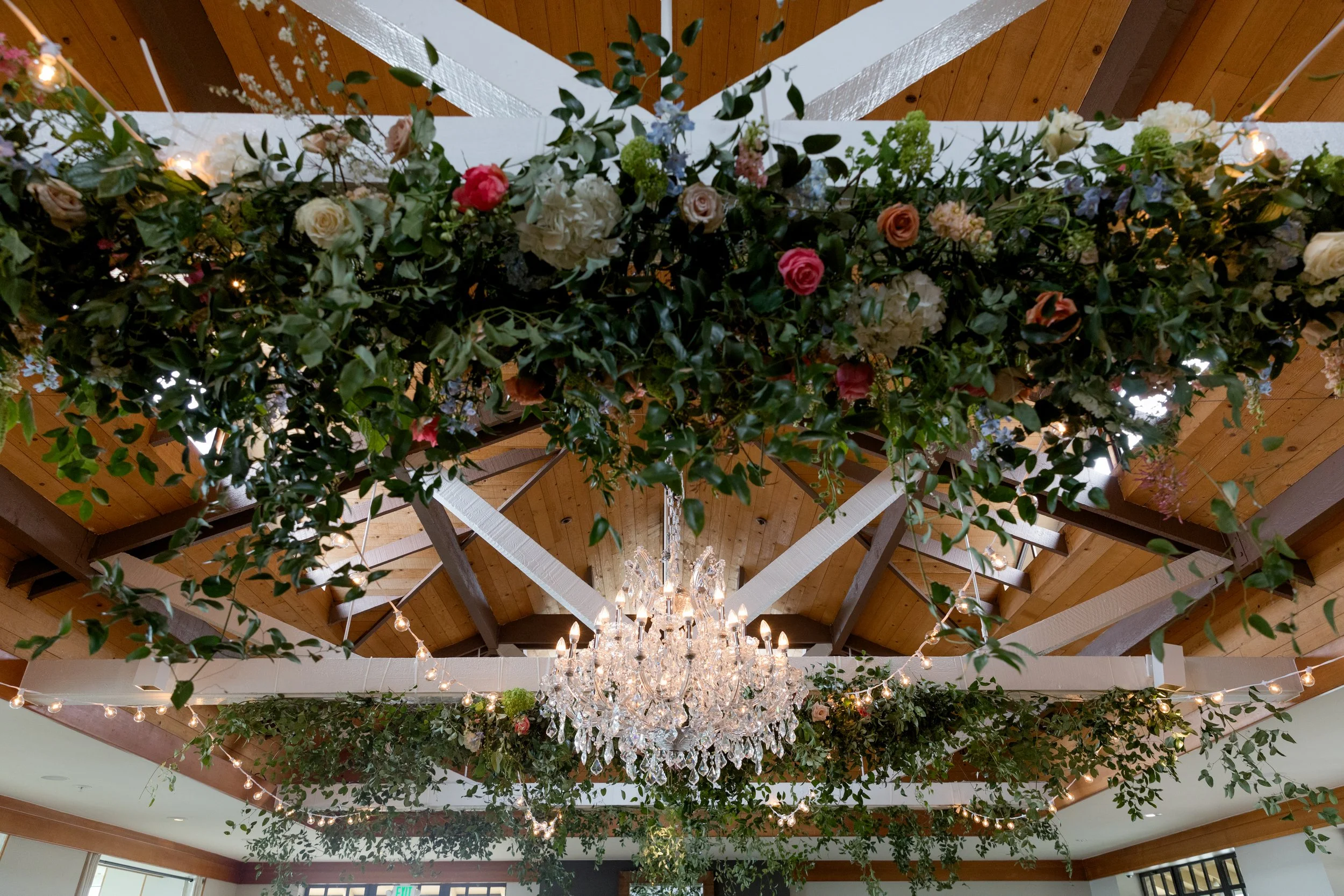 Elegant wedding reception venue decorated with lush greenery, flowers, string lights, and a crystal chandelier