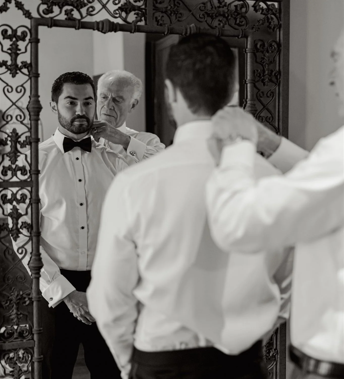 A black-and-white photo captures a young groom in a white tuxedo shirt and bow tie receiving help adjusting his collar from his father.