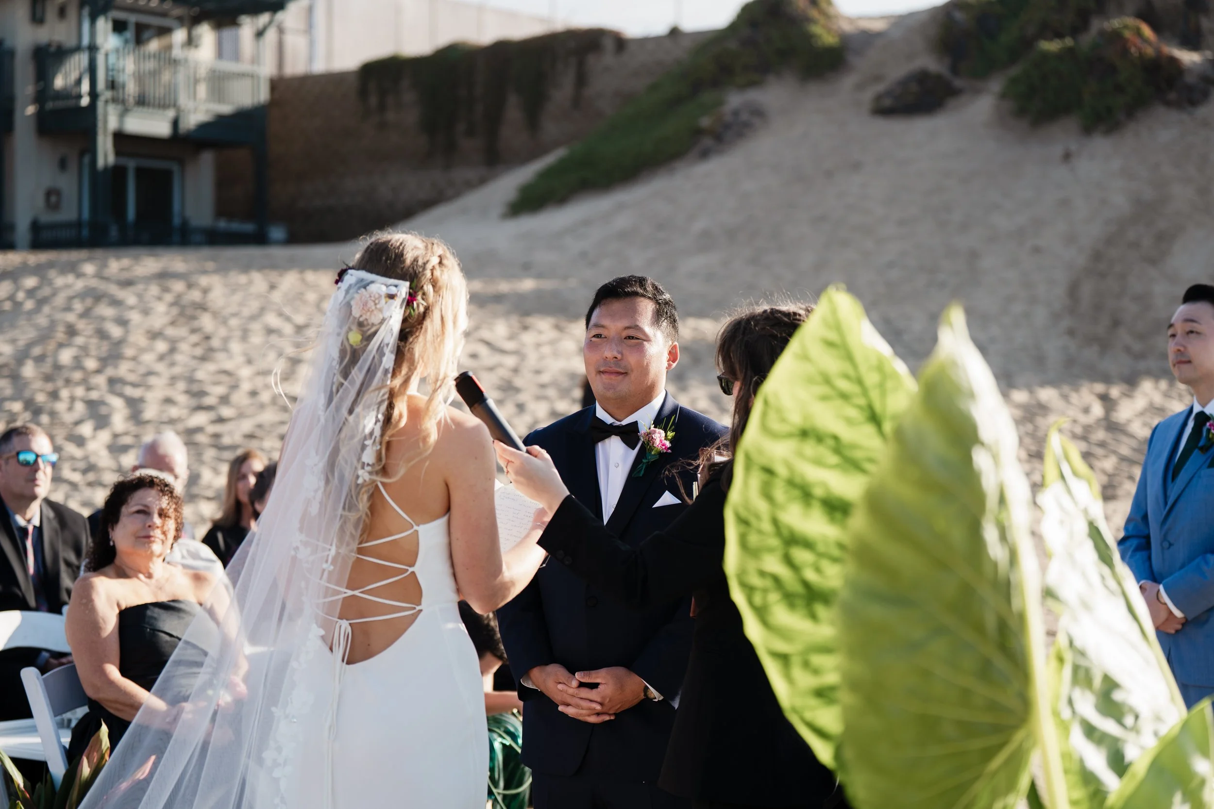 Bride reading vows to groom during an intimate beach wedding ceremony with guests seated on the sand and tropical greenery in the foreground.