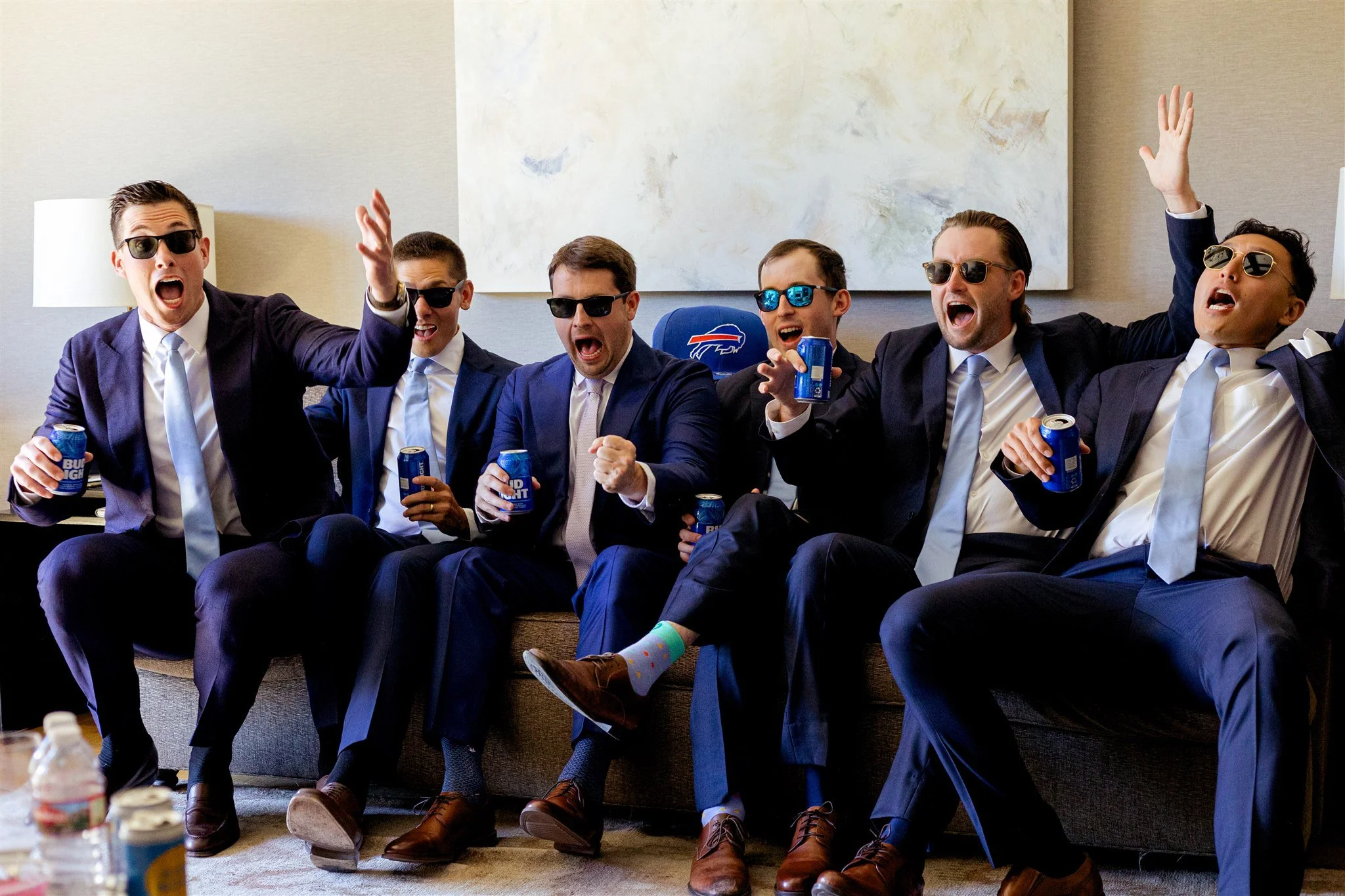 Six suited men in sunglasses sit on a couch, cheering with beers while getting ready for a wedding.