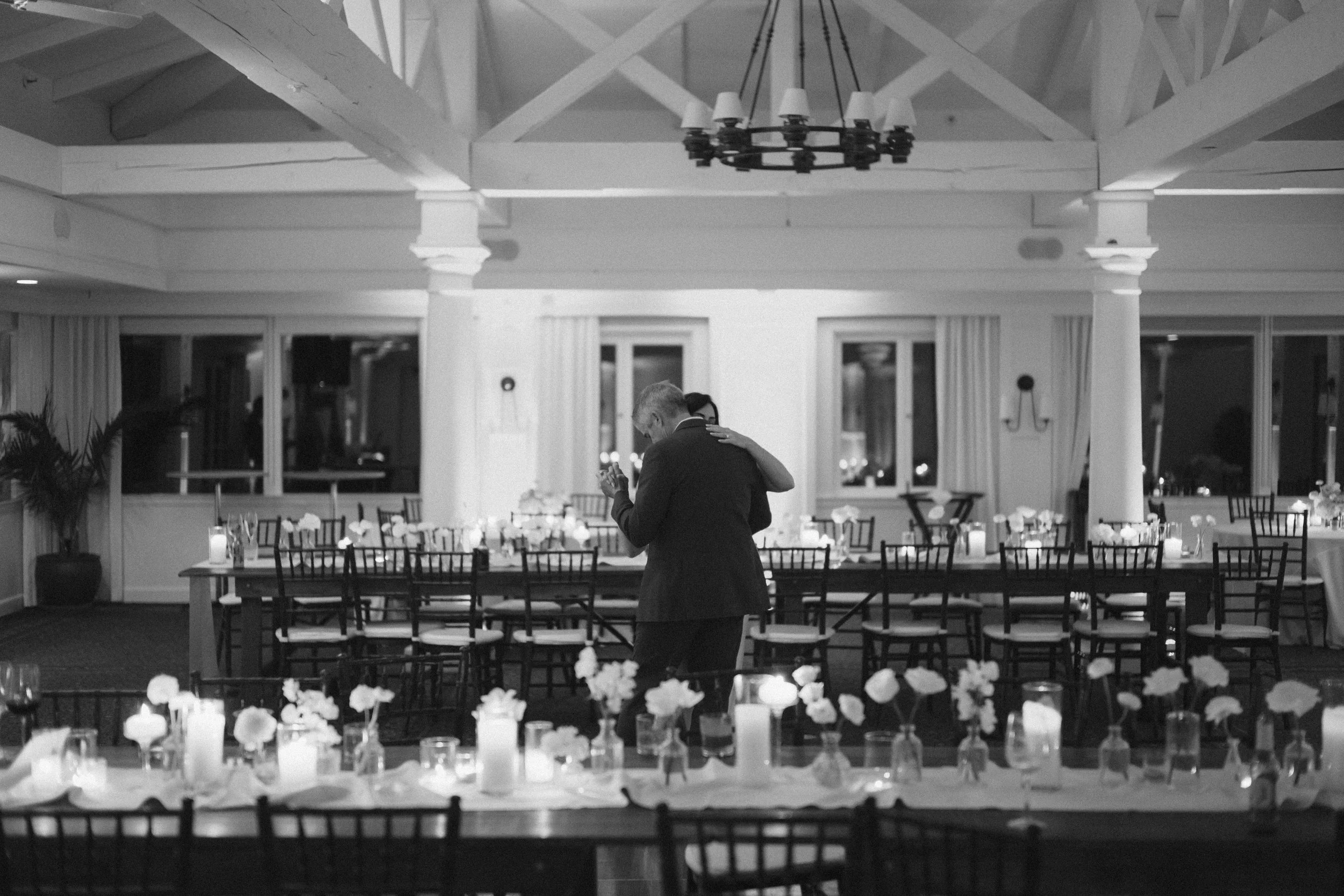 Black-and-white photo of a couple slow dancing alone in a softly lit wedding reception hall, surrounded by long tables set with candles, glassware, and floral arrangements beneath exposed white beams and a chandelier.