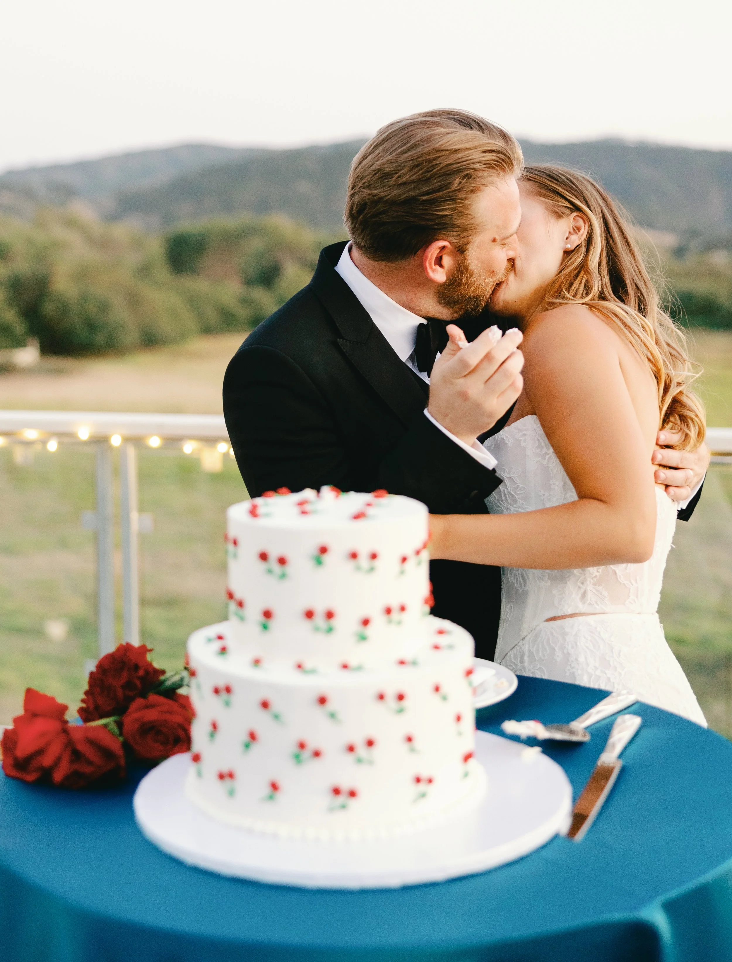 Newlyweds kiss behind their two-tiered white wedding cake decorated with hand-painted red roses at an outdoor reception.
