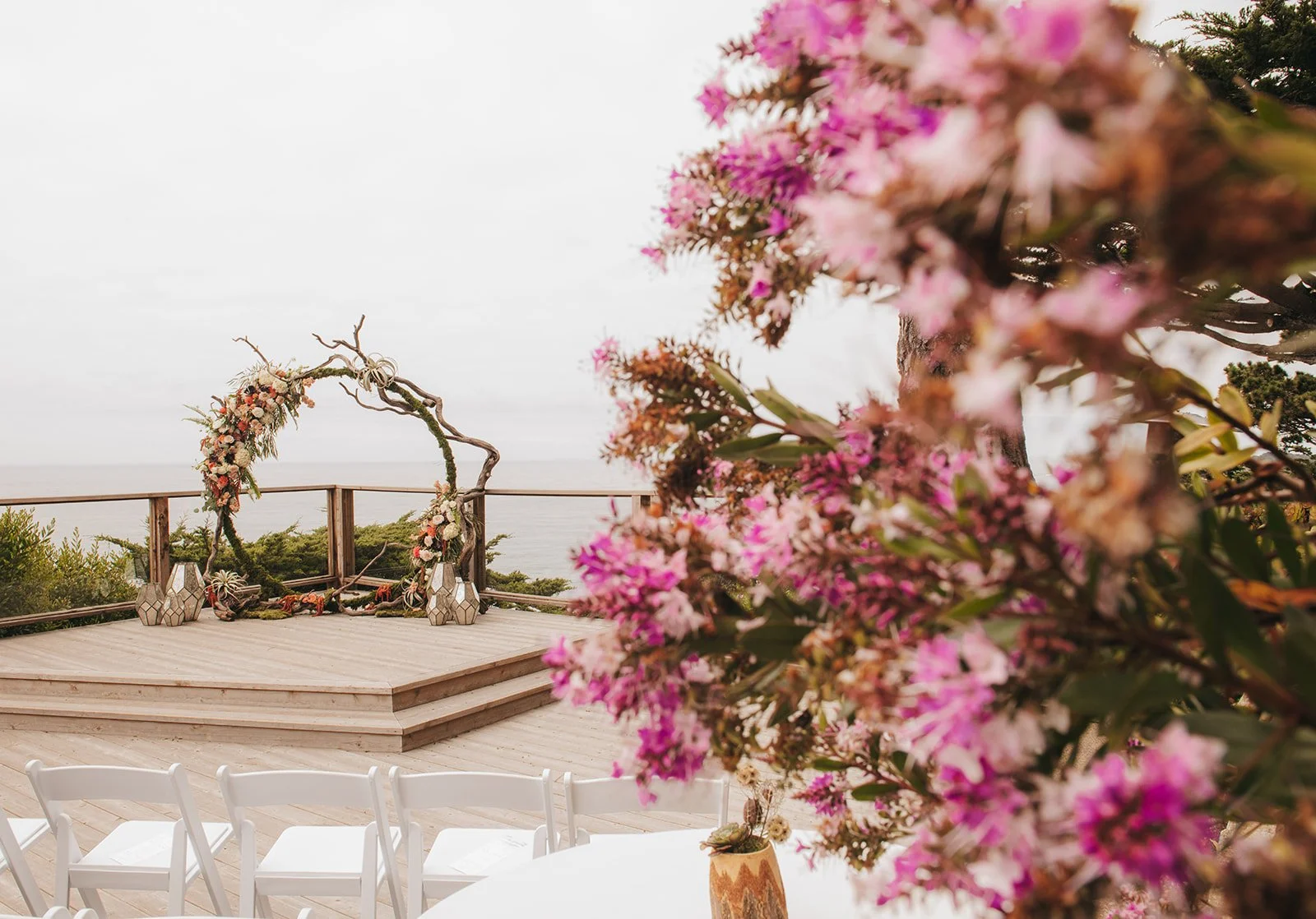 Coastal wedding ceremony setup on a wooden deck overlooking the ocean, featuring a rustic branch arch adorned with floral arrangements and vibrant pink flowers in the foreground.