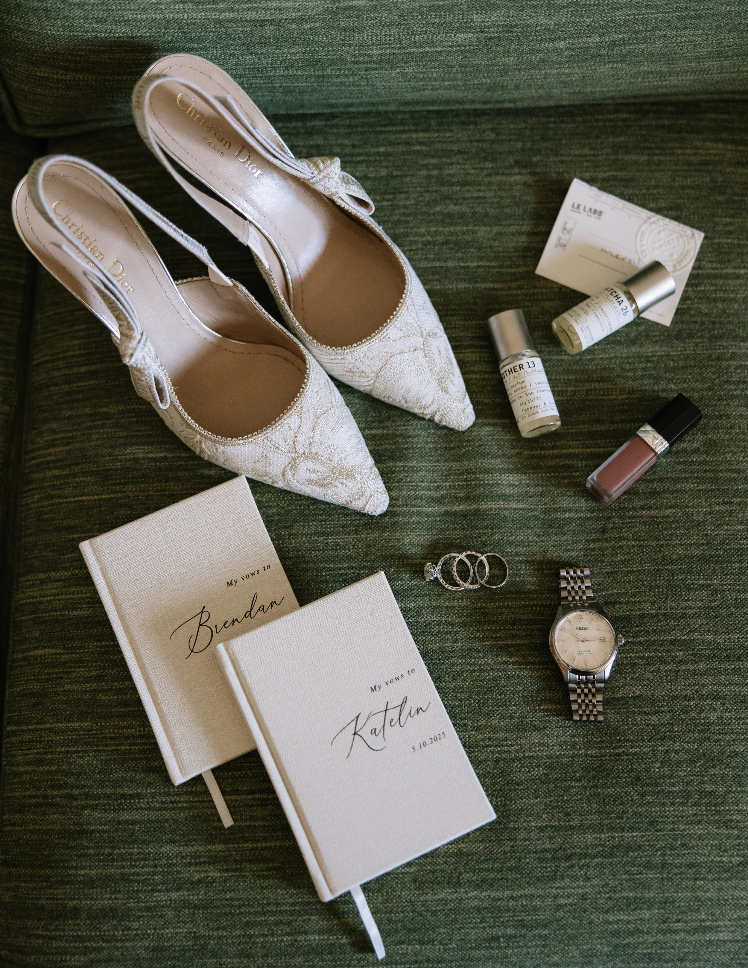 Flat lay of bridal details on a green textured sofa, featuring lace slingback heels by Christian Dior, personalized vow books, wedding rings, a silver Seiko watch, and fragrances from Le Labo.