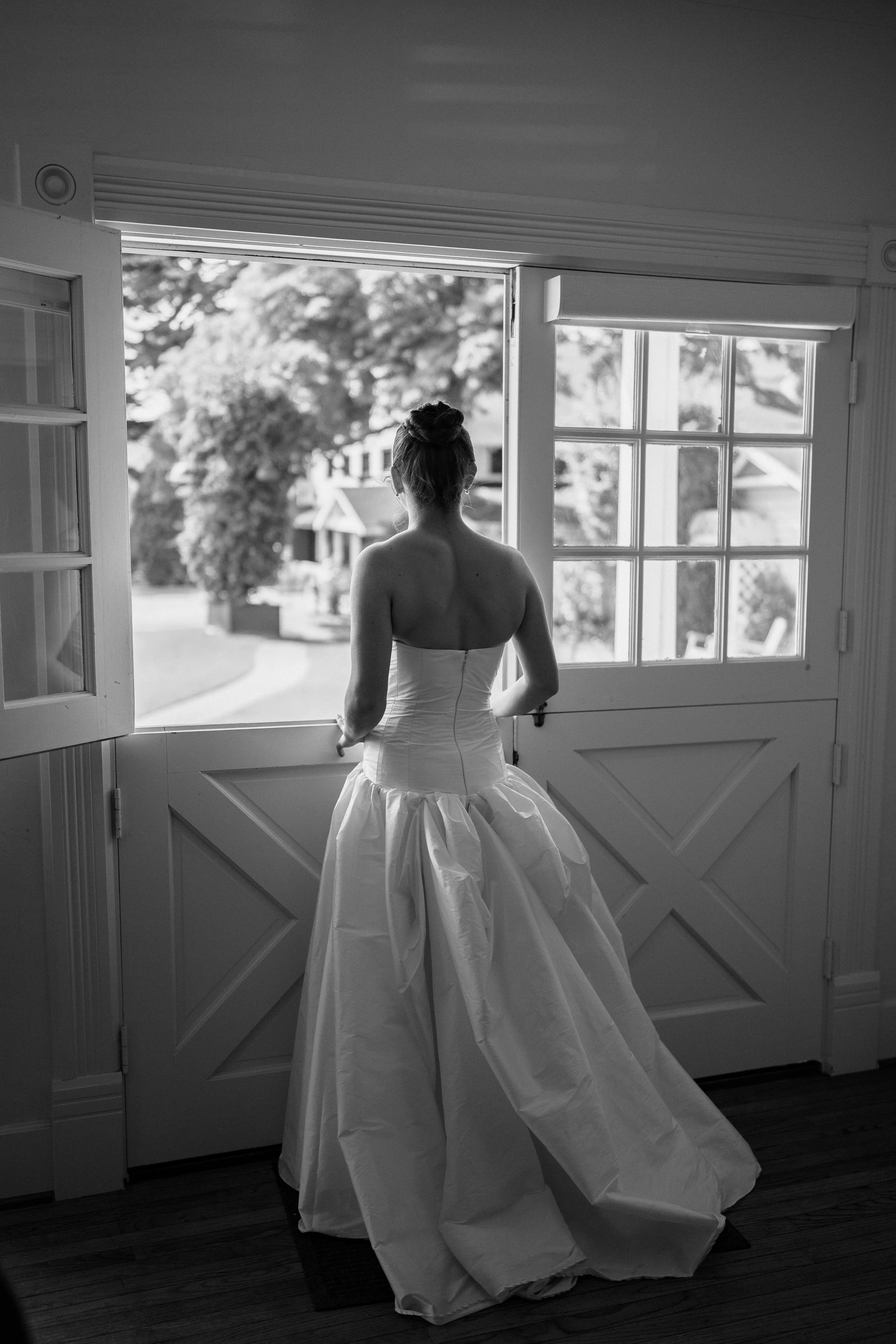 A bride in a strapless wedding gown stands with her back to the camera, looking out through open barn-style doors in a black-and-white portrait.