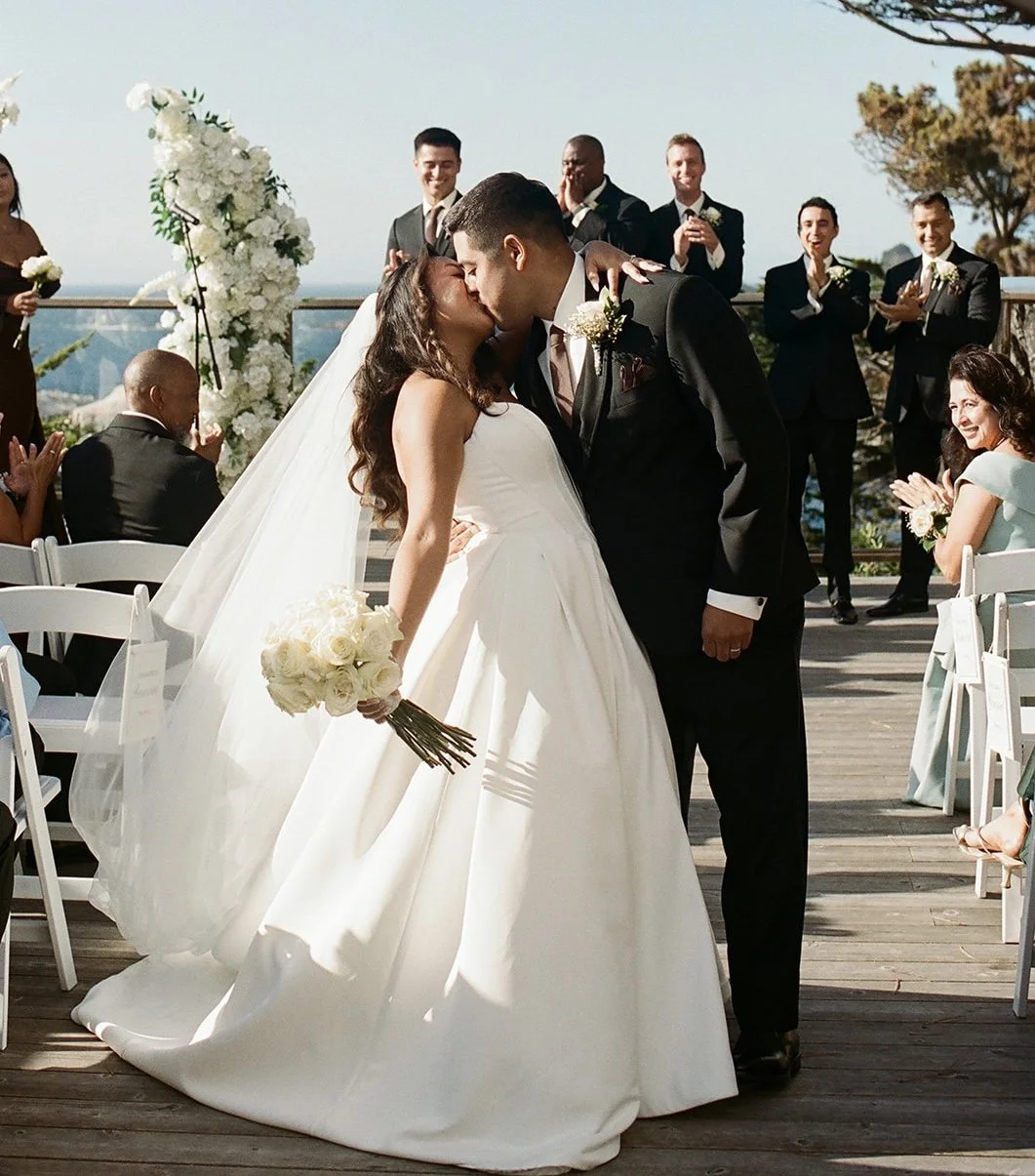 Bride and groom kiss at an outdoor wedding ceremony as guests applaud.