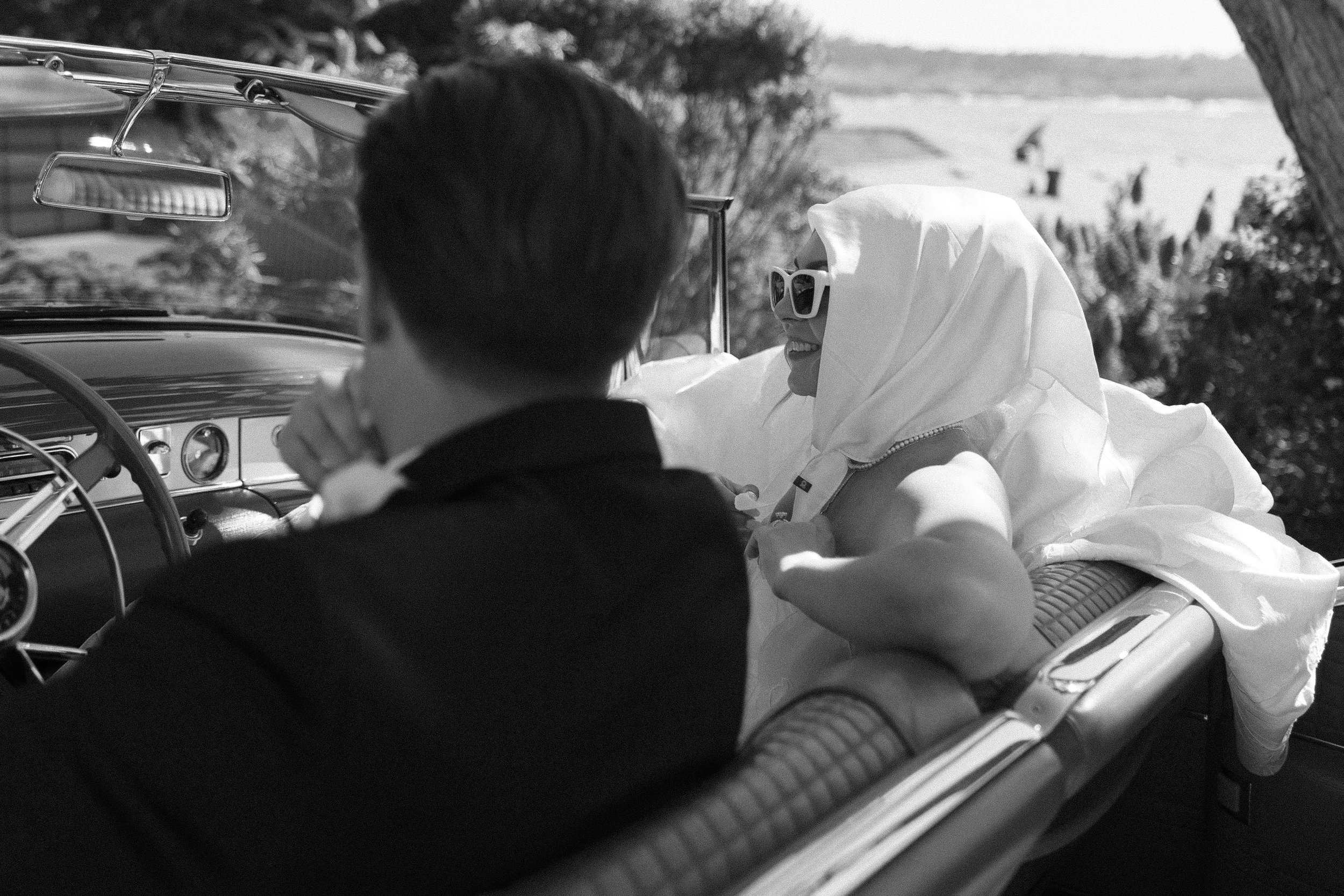 Black-and-white photo of a bride in a headscarf and white sunglasses smiling from the back seat of a vintage convertible, with the groom in a tuxedo seated in front and a coastal landscape in the background.