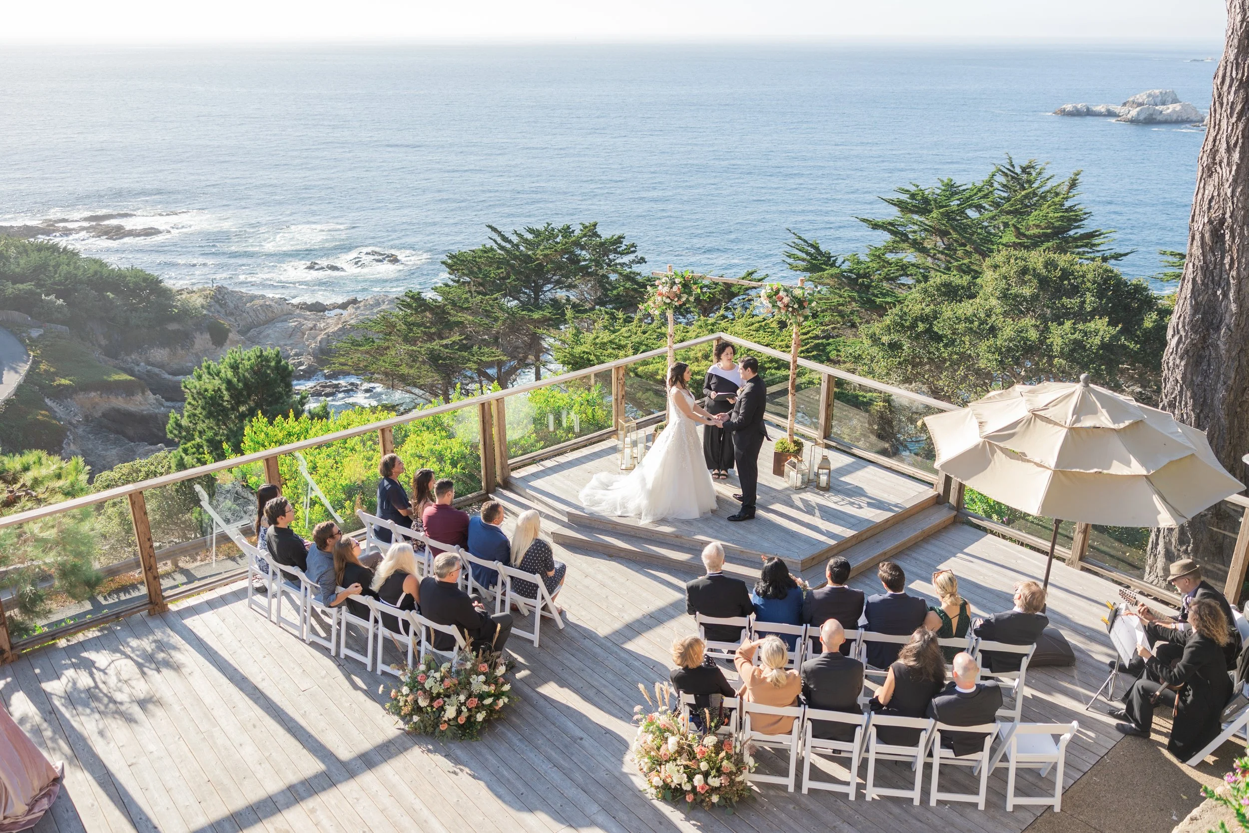Outdoor wedding ceremony on a coastal deck overlooking the Pacific Ocean, surrounded by guests and floral arrangements