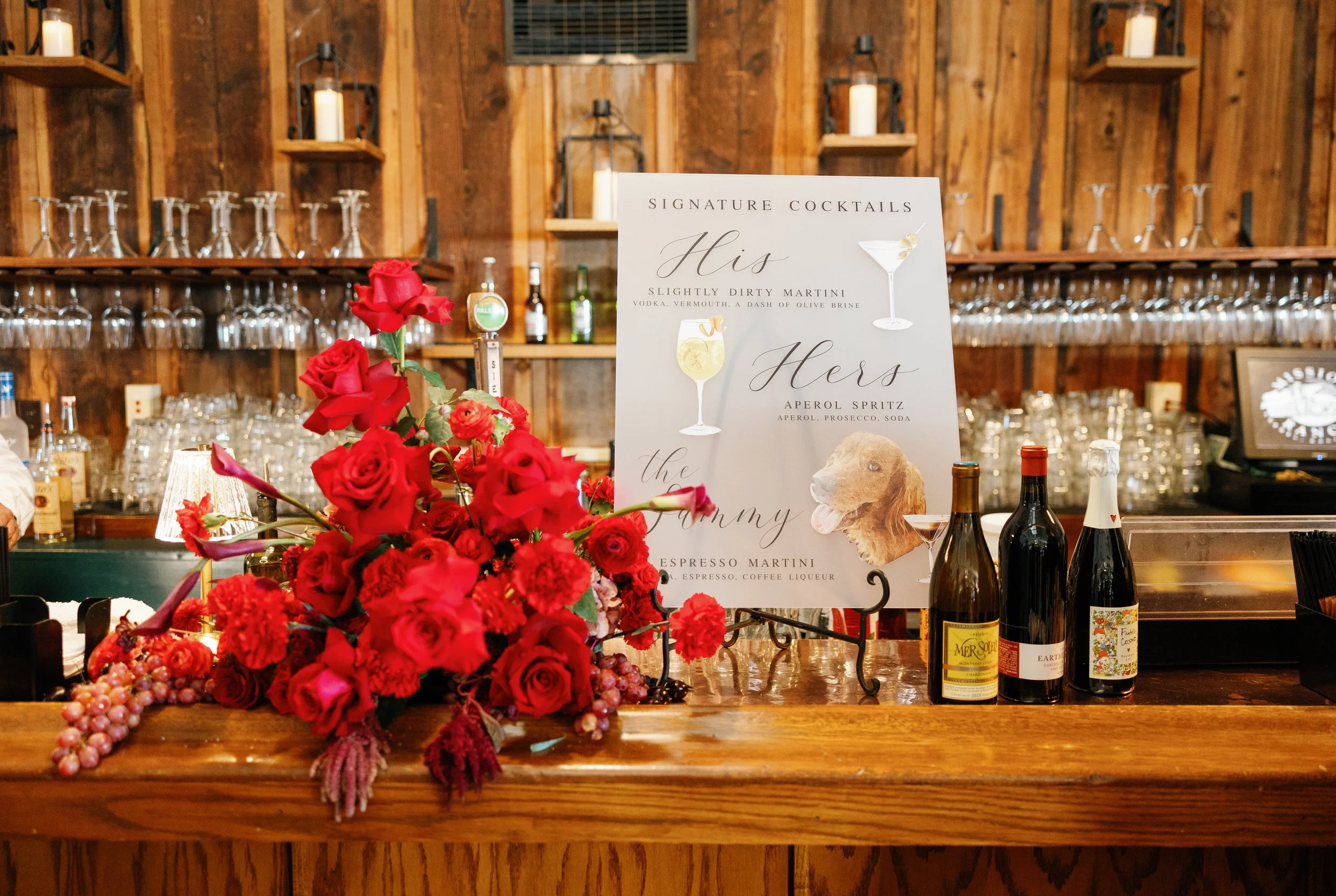 Signature cocktails sign on a rustic wooden bar, decorated with a lush red rose and grape centerpiece, next to bottles of wine and champagne.