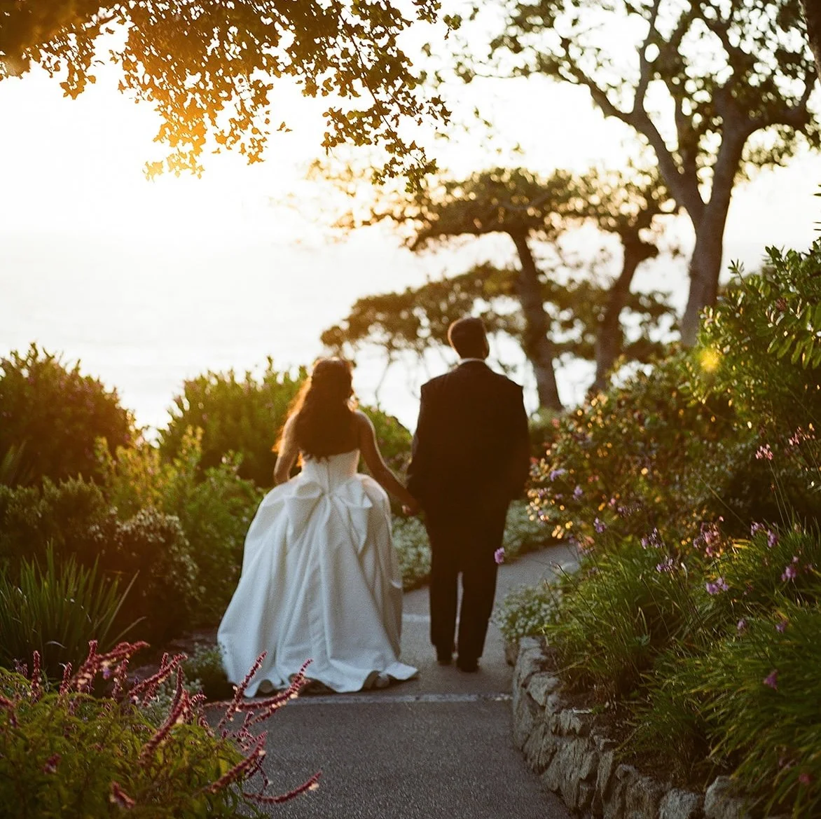 Bride and groom walk hand-in-hand down a garden path at sunset, with a water view in the background.