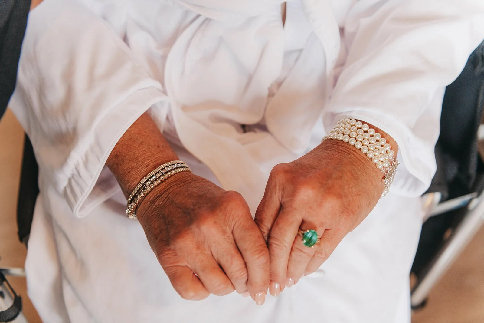 Close-up of a wedding guest’s hands adorned with pearl bracelets, gold jewelry, and a green gemstone ring, paired with an elegant white outfit.