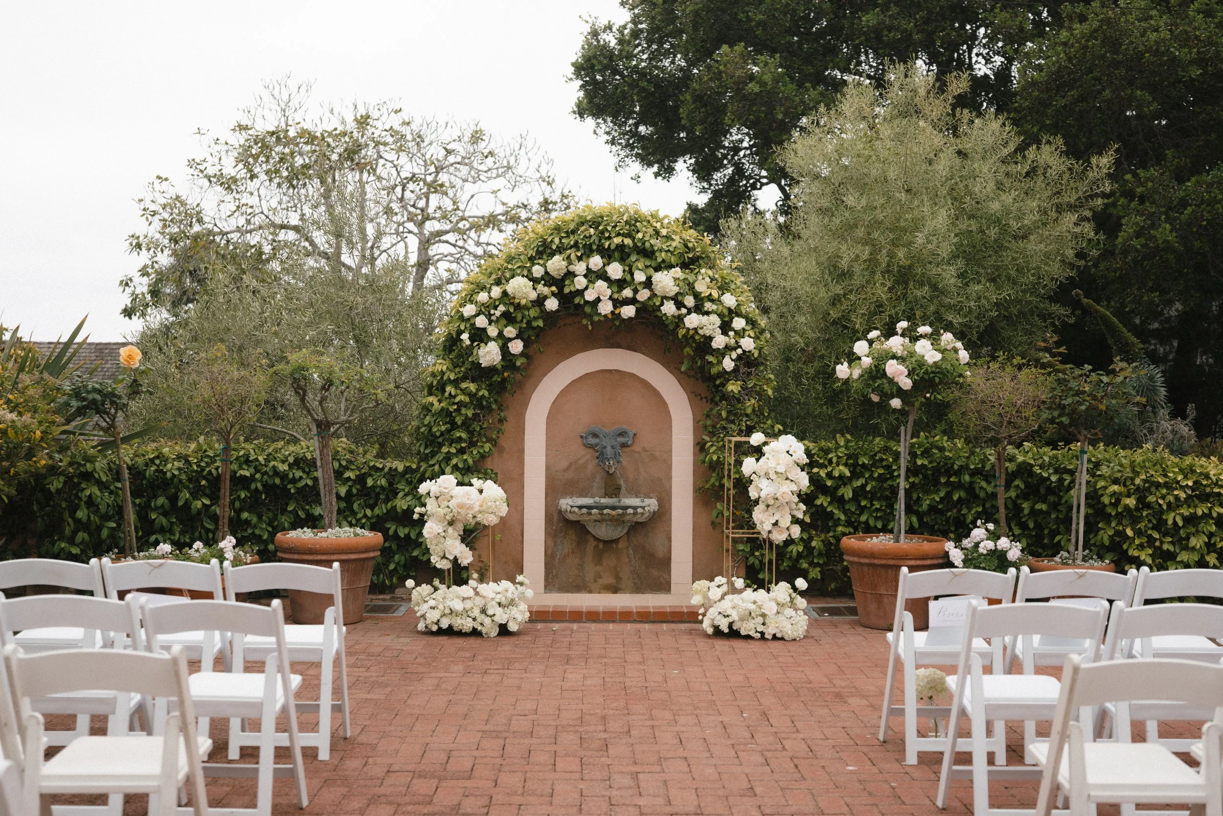 Outdoor wedding ceremony setup with white folding chairs arranged on a brick patio facing a small stone fountain set into an arched wall, surrounded by lush greenery and white floral arrangements, including roses and climbing vines.