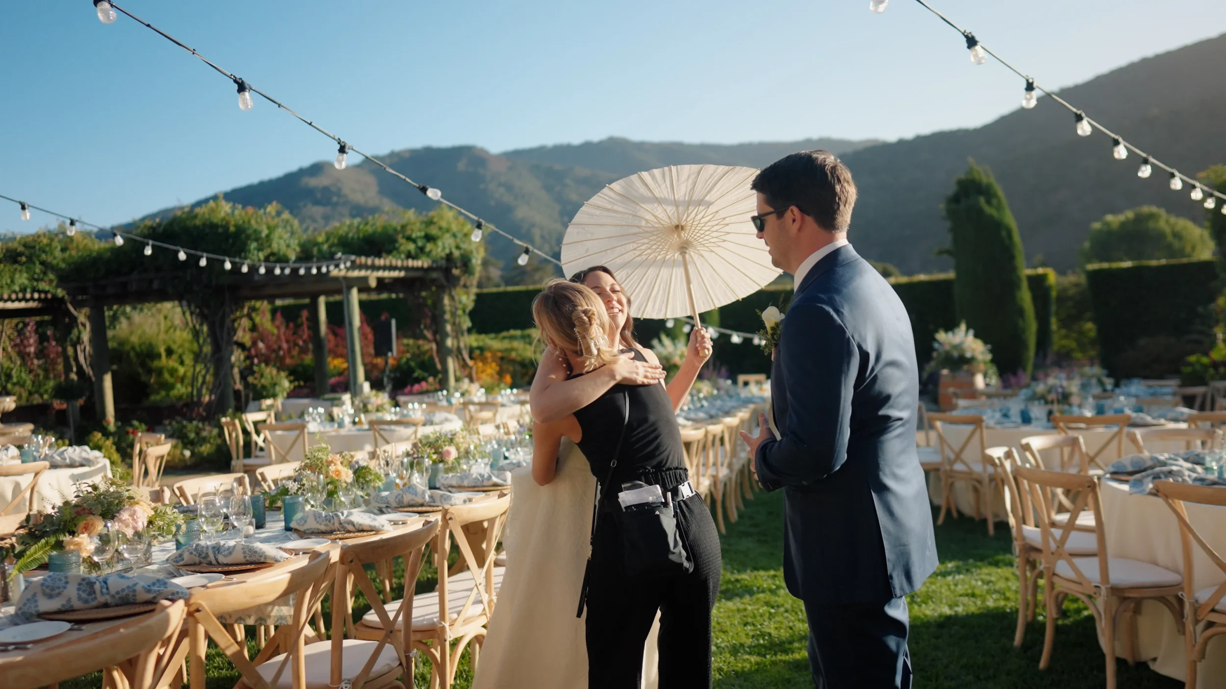 A bride hugs her wedding planner, Jessica, while holding a white parasol at an outdoor reception, with the groom standing nearby; long tables are set with flowers and tableware under string lights, surrounded by greenery and hills in the background.