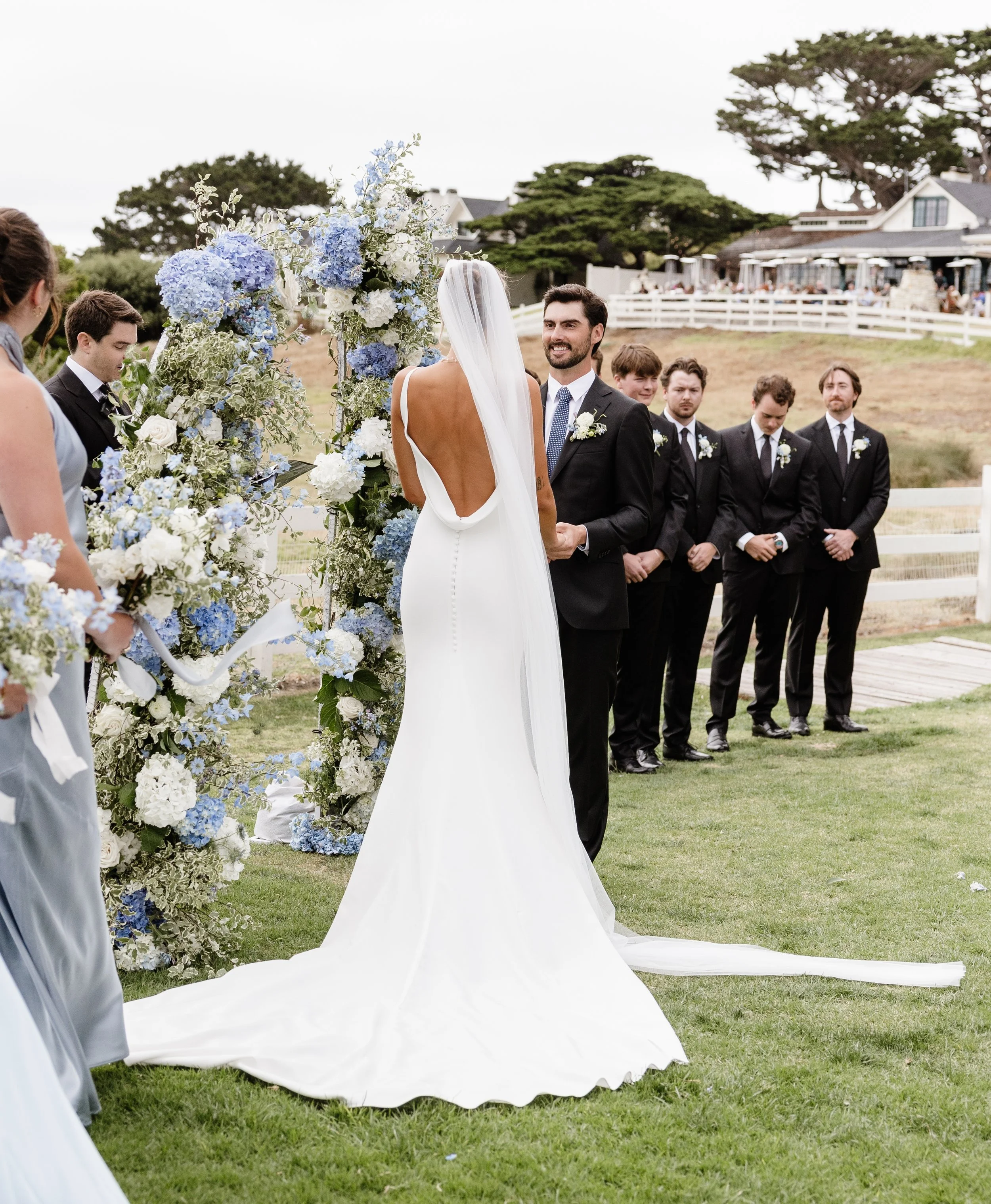 Bride in a minimalist low-back wedding dress stands at a blue and white floral ceremony arch facing groom during an outdoor wedding ceremony.
