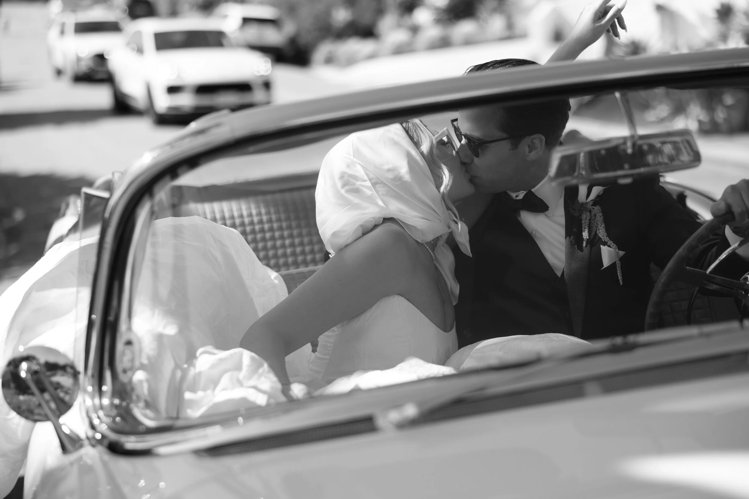 Black-and-white photo of a newlywed couple sharing a kiss in a vintage convertible, the bride wearing a headscarf and gown while the groom, in a tuxedo and sunglasses, sits in the driver’s seat.