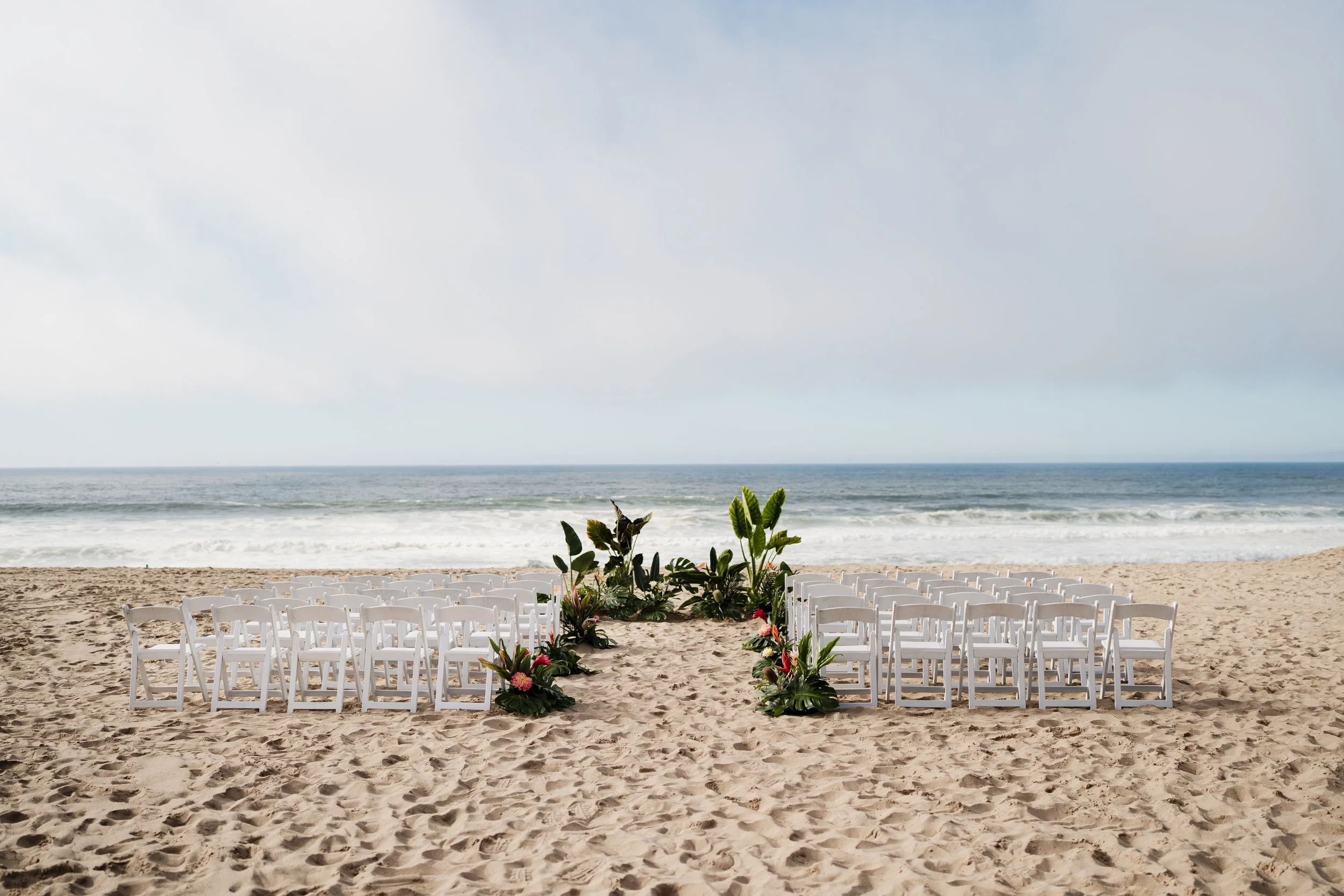 Beach wedding ceremony setup with rows of white chairs facing the ocean and tropical floral arrangements lining the aisle.