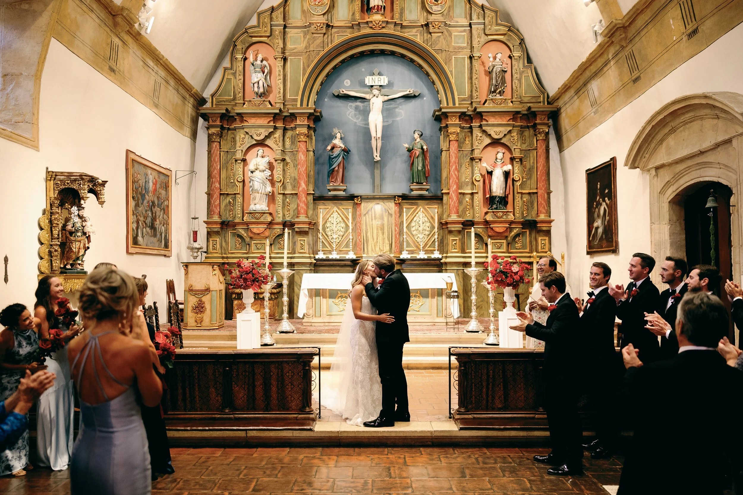 Newlywed couple sharing a kiss at the altar inside Carmel Mission Basilica surrounded by wedding guests and historic Spanish architecture.