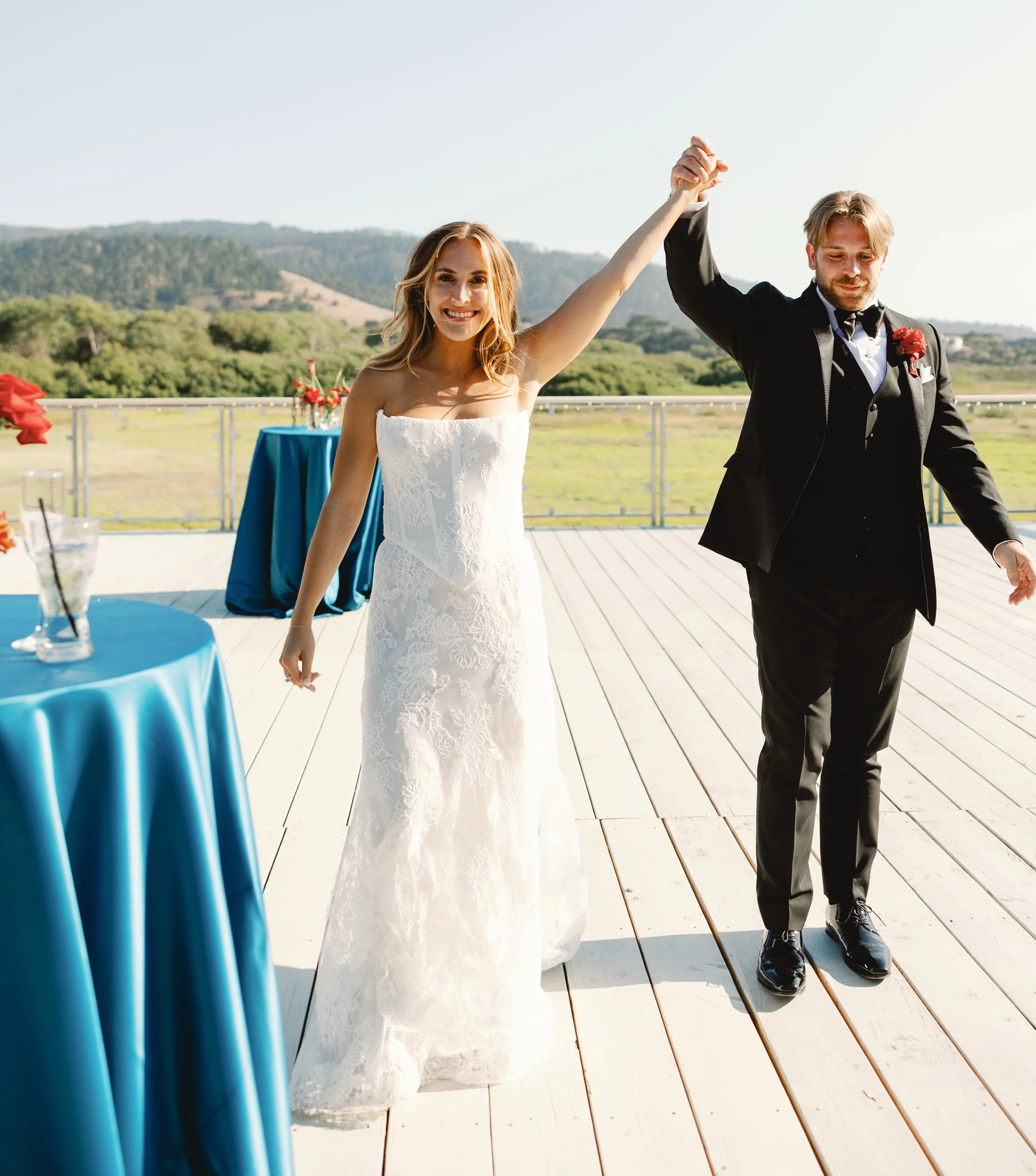 Bride and groom raise their joined hands in a joyful grand entrance onto an outdoor deck with rolling hills behind them.