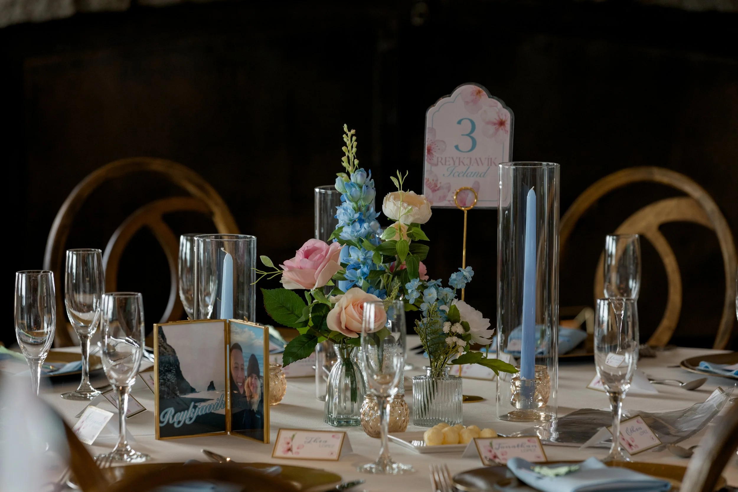 Wedding table setting for table 3, 'Reykjavík, Iceland,' with blue taper candles, pink and peach roses, blue delphinium, and a framed travel photo of a couple.