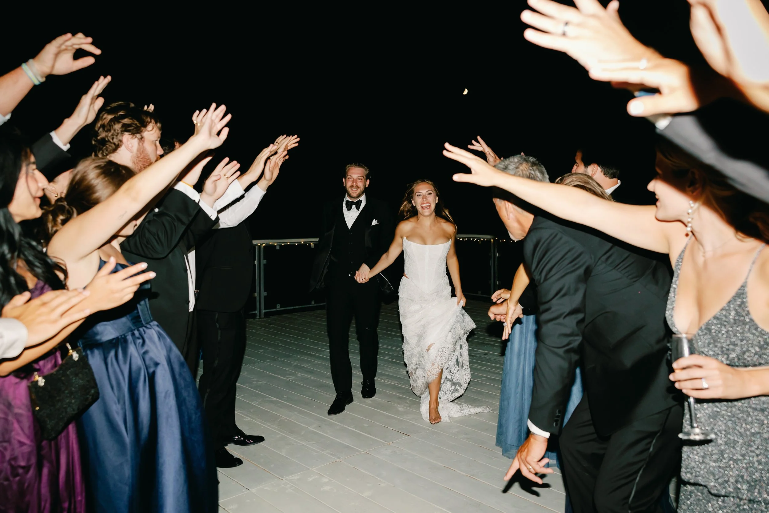 Bride and groom holding hands and laughing as they walk through a cheering crowd of wedding guests with raised hands, on an outdoor deck at night.