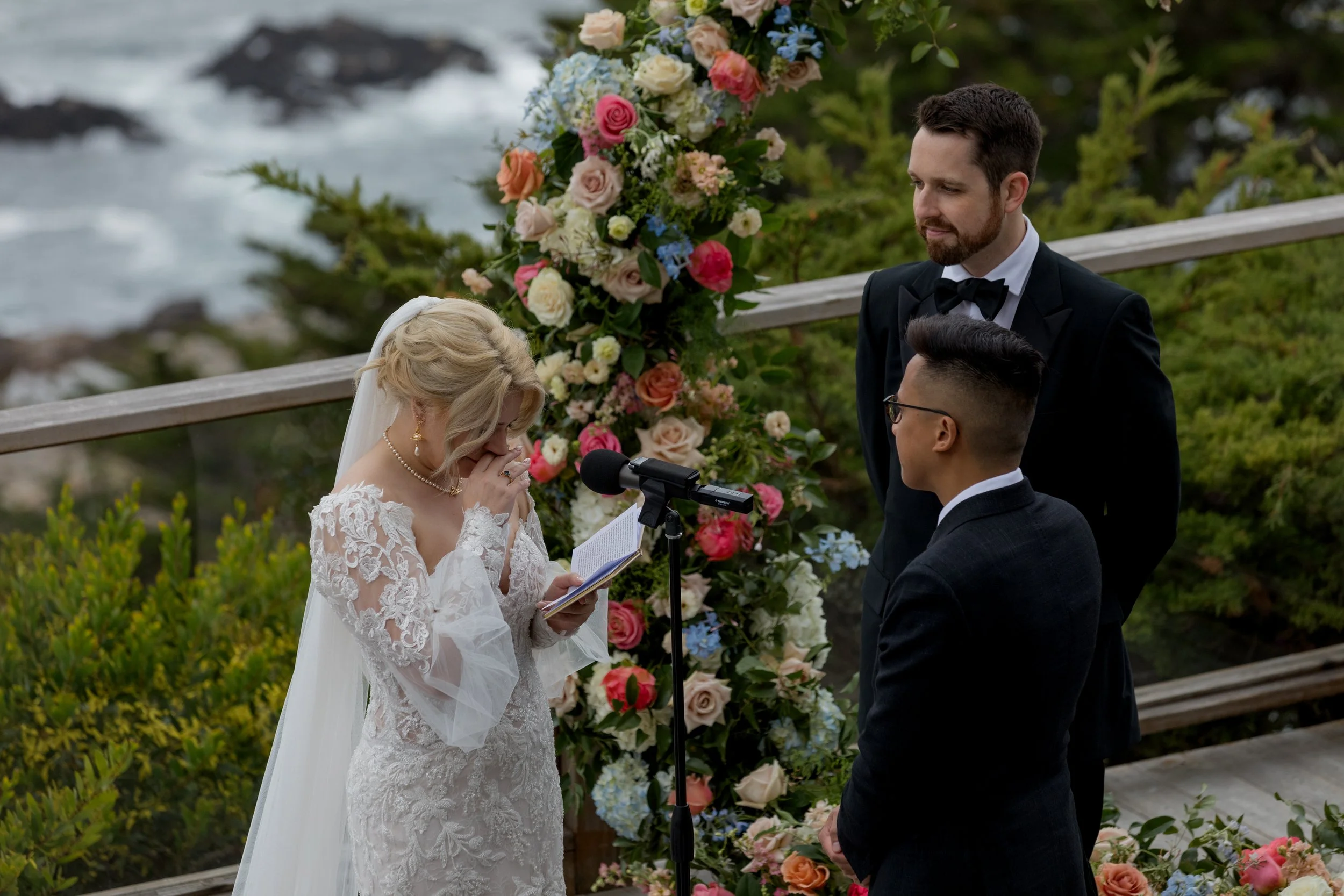 Bride reading wedding vows to groom during an oceanfront ceremony surrounded by colorful flowers