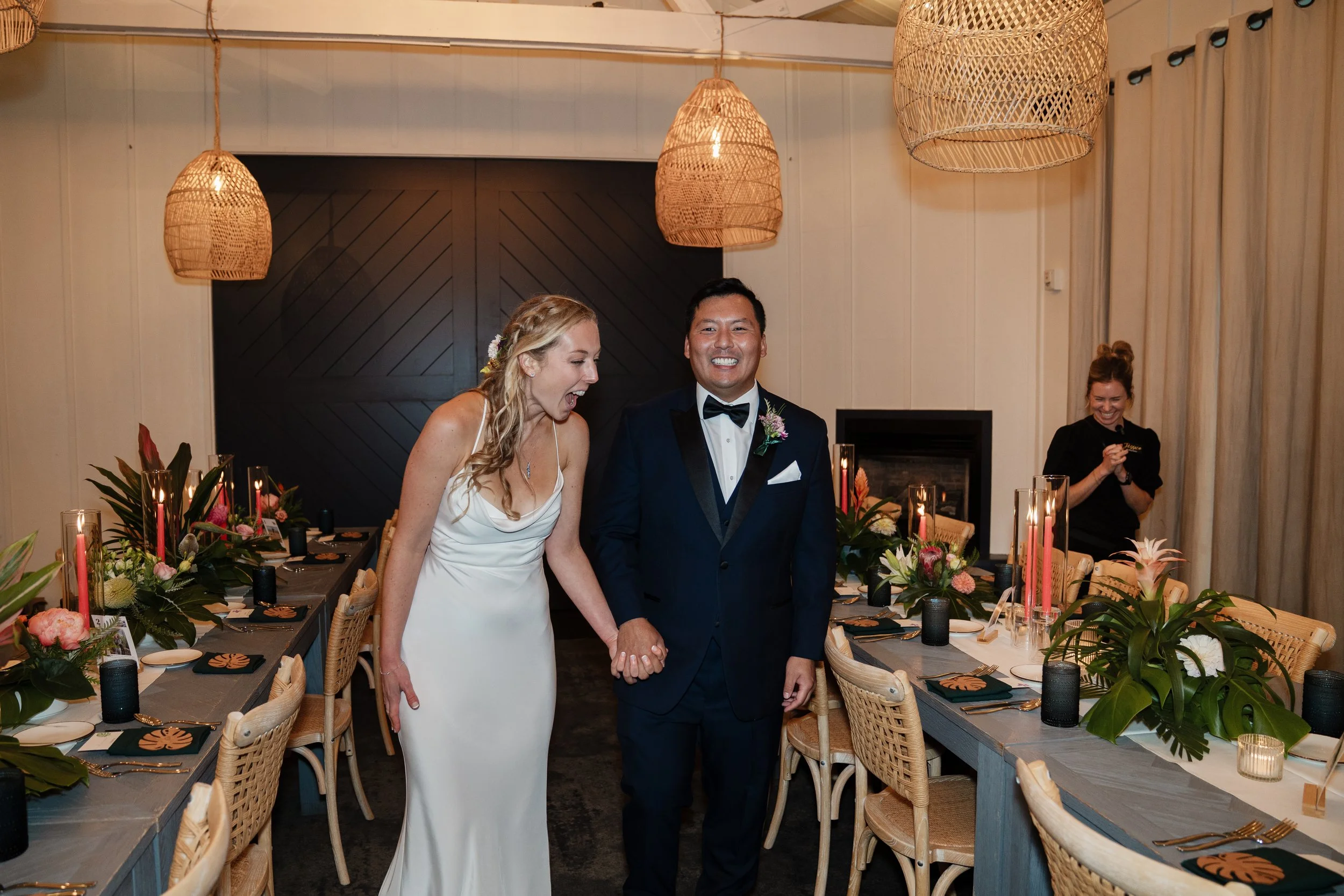 Bride and groom smiling and holding hands as they make their reception entrance into an elegant dining room with tropical floral arrangements and woven pendant lights.