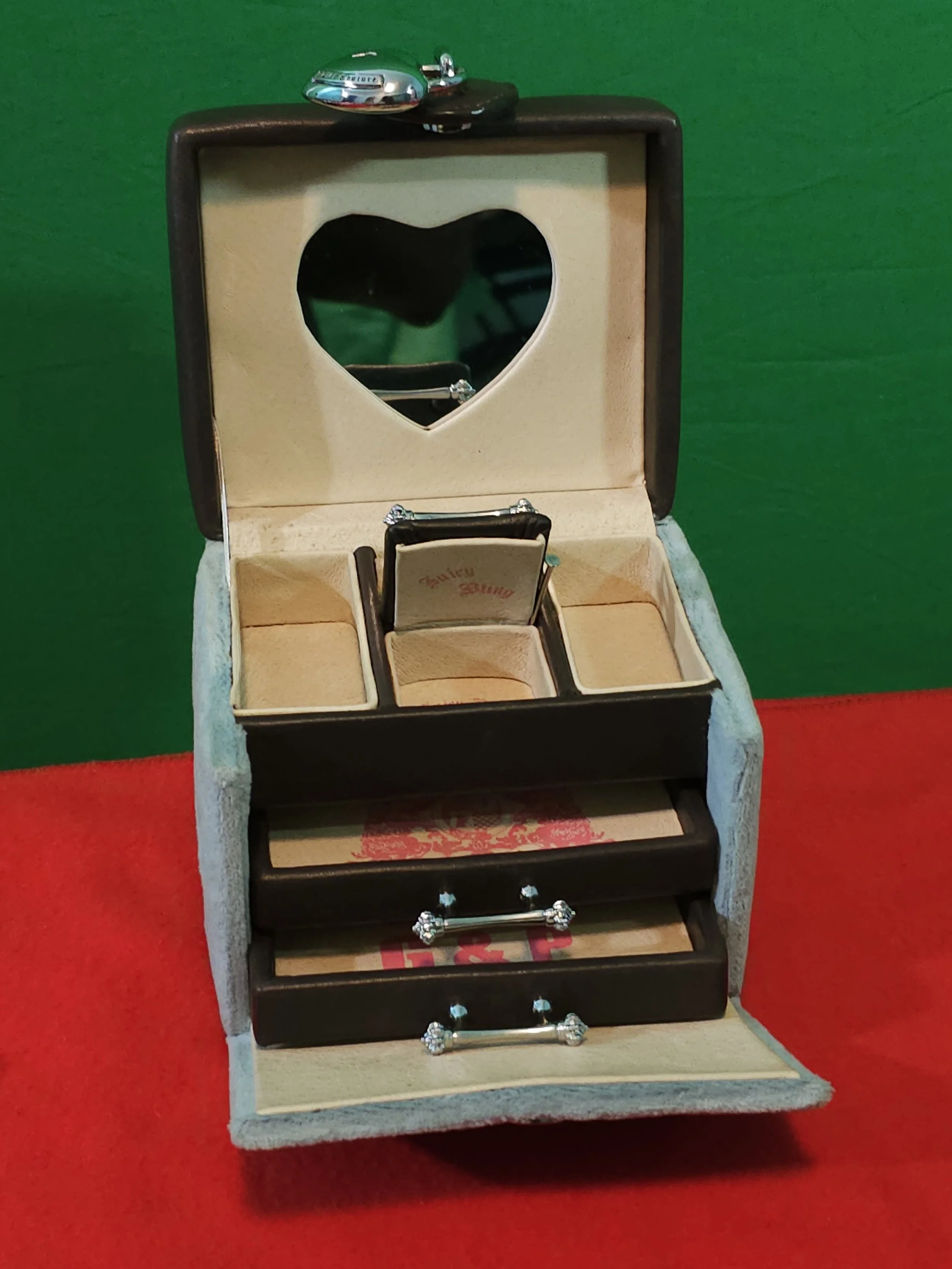 A small vintage jewelry box with a heart-shaped mirror on the inside of the lid, holding a ring with a diamond. The box has multiple compartments and drawers, decorated with floral patterns and silver handles.