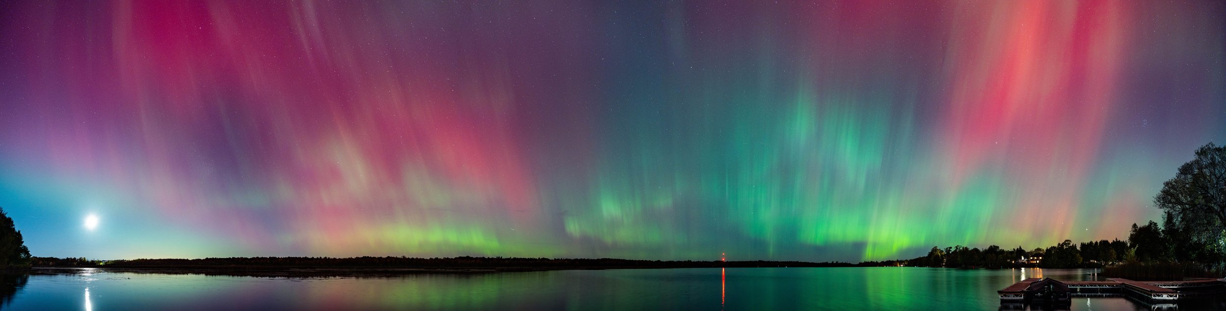 Northern lights (aurora borealis) display over a body of water at night with a clear sky, some trees, and a faint moon visible.