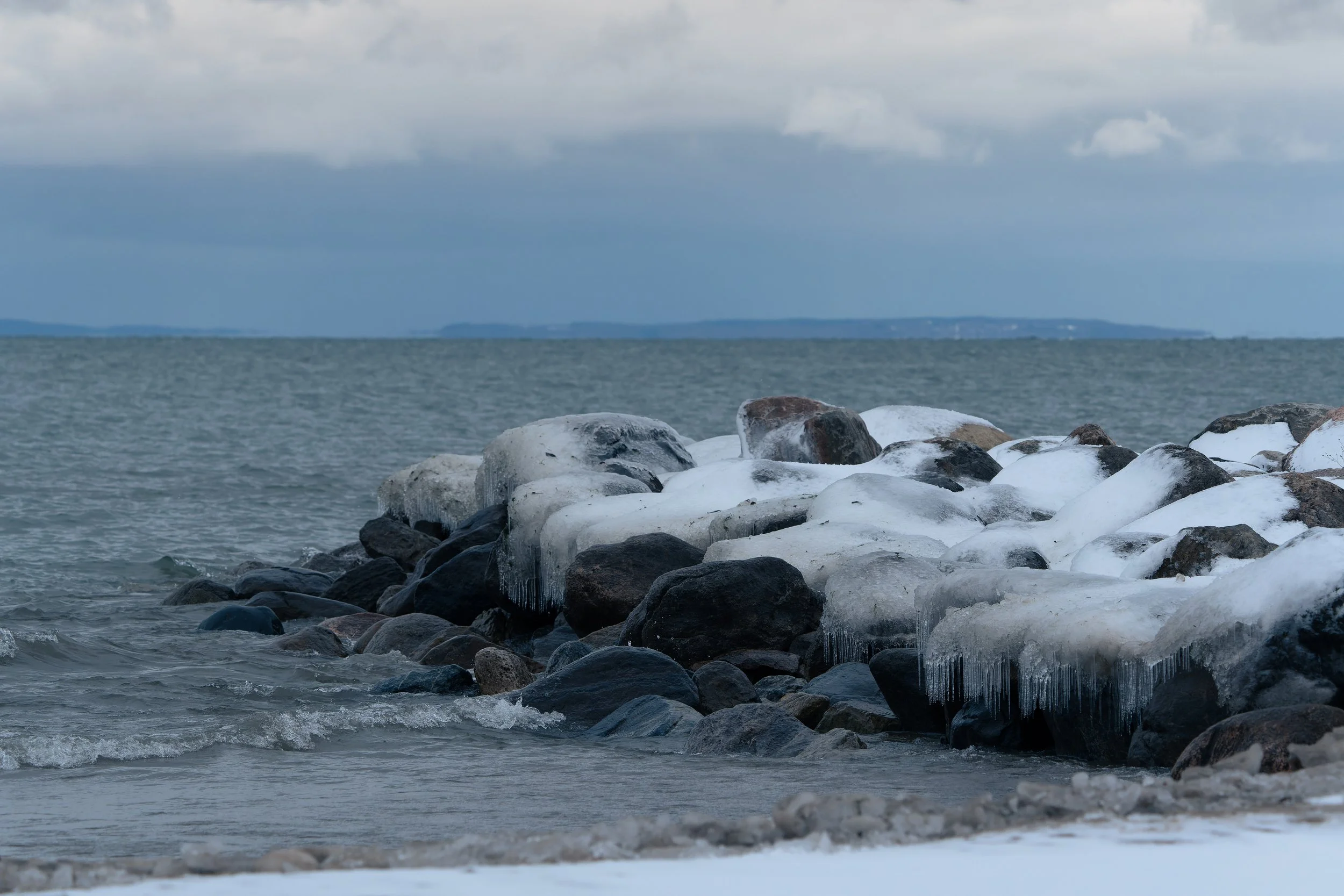 A shoreline with large rocks and ice, with a body of water and a cloudy sky in the background.