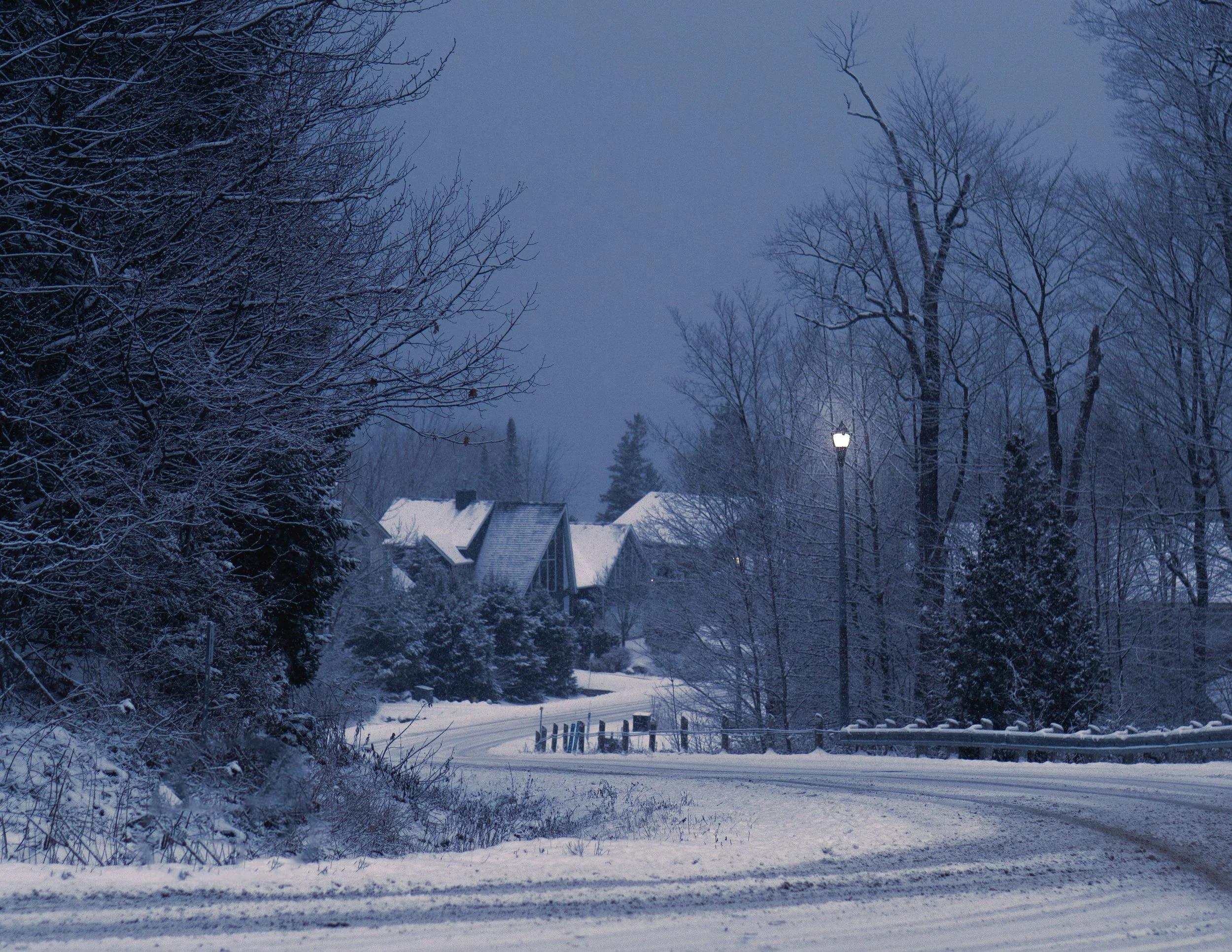 Snow-covered residential neighborhood with rooftops, bare trees, snow on the ground, streetlights, and a cloudy evening sky.