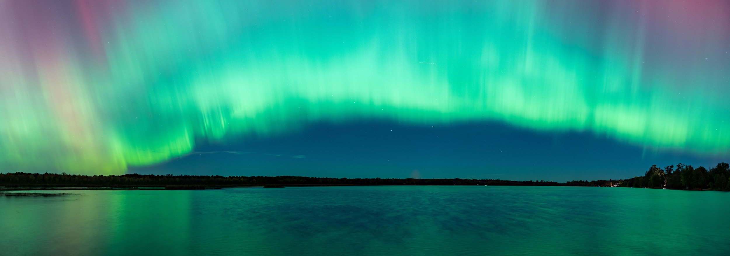 Northern lights over a body of water at night with a silhouette of trees in the distance.