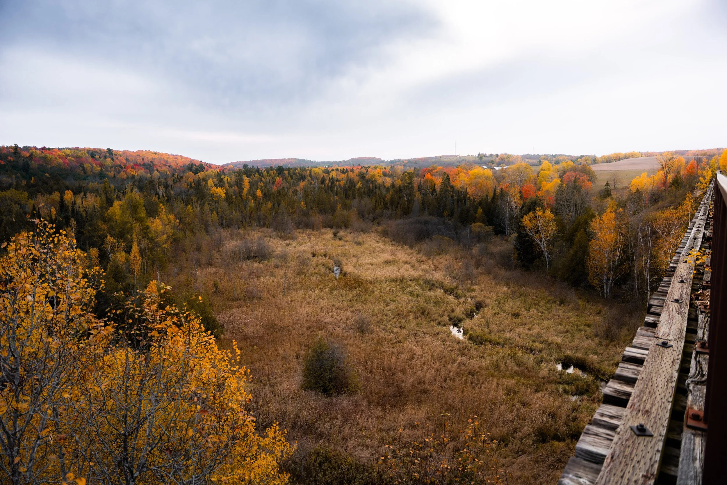 Orange Corners Trestle Bridge - A scenic view of a hilly landscape with trees displaying fall colors, a cloudy sky, and a section of a wooden train track on the right side of the image.