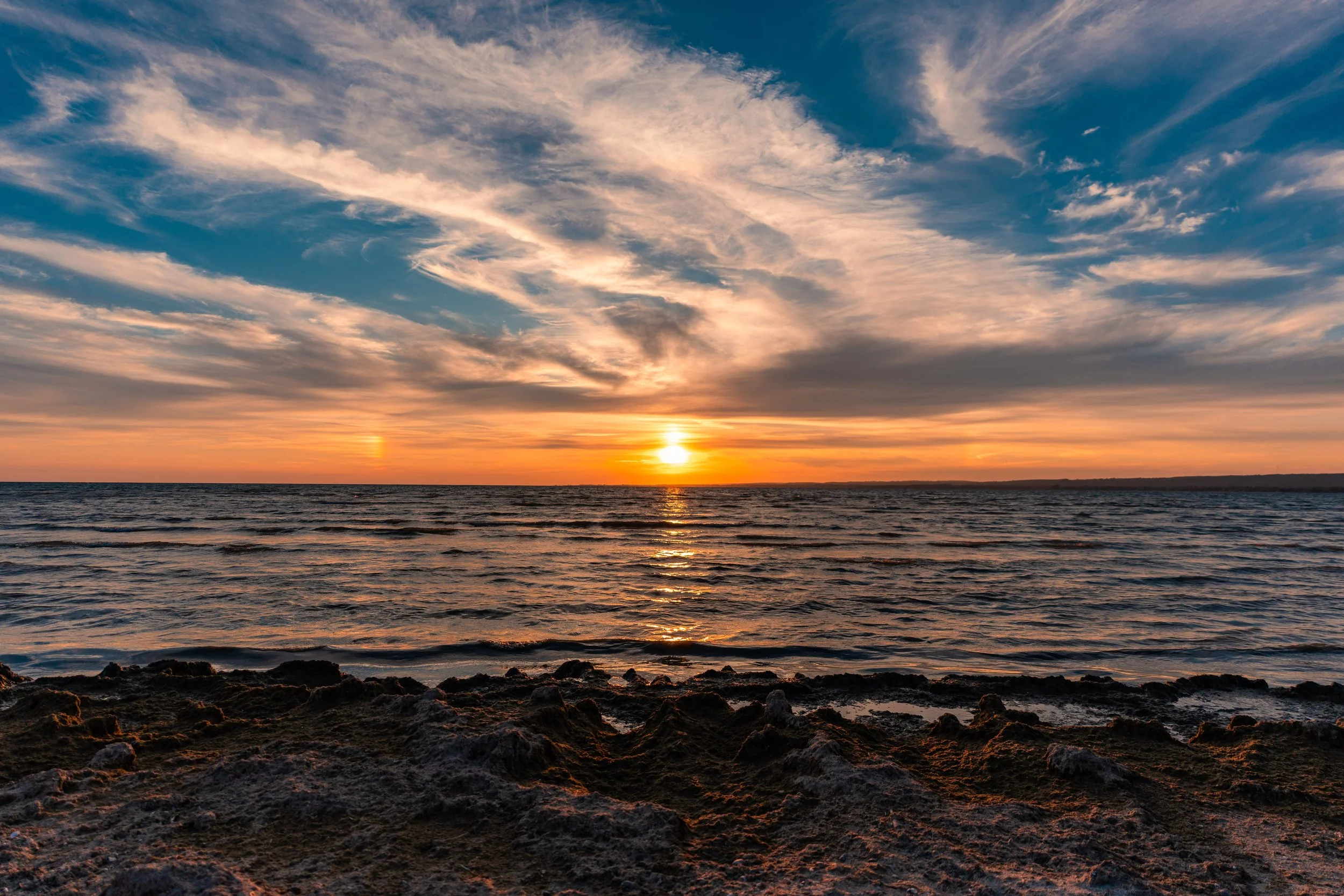 Sunset over the ocean with clouds in the sky and rocky shoreline in the foreground.