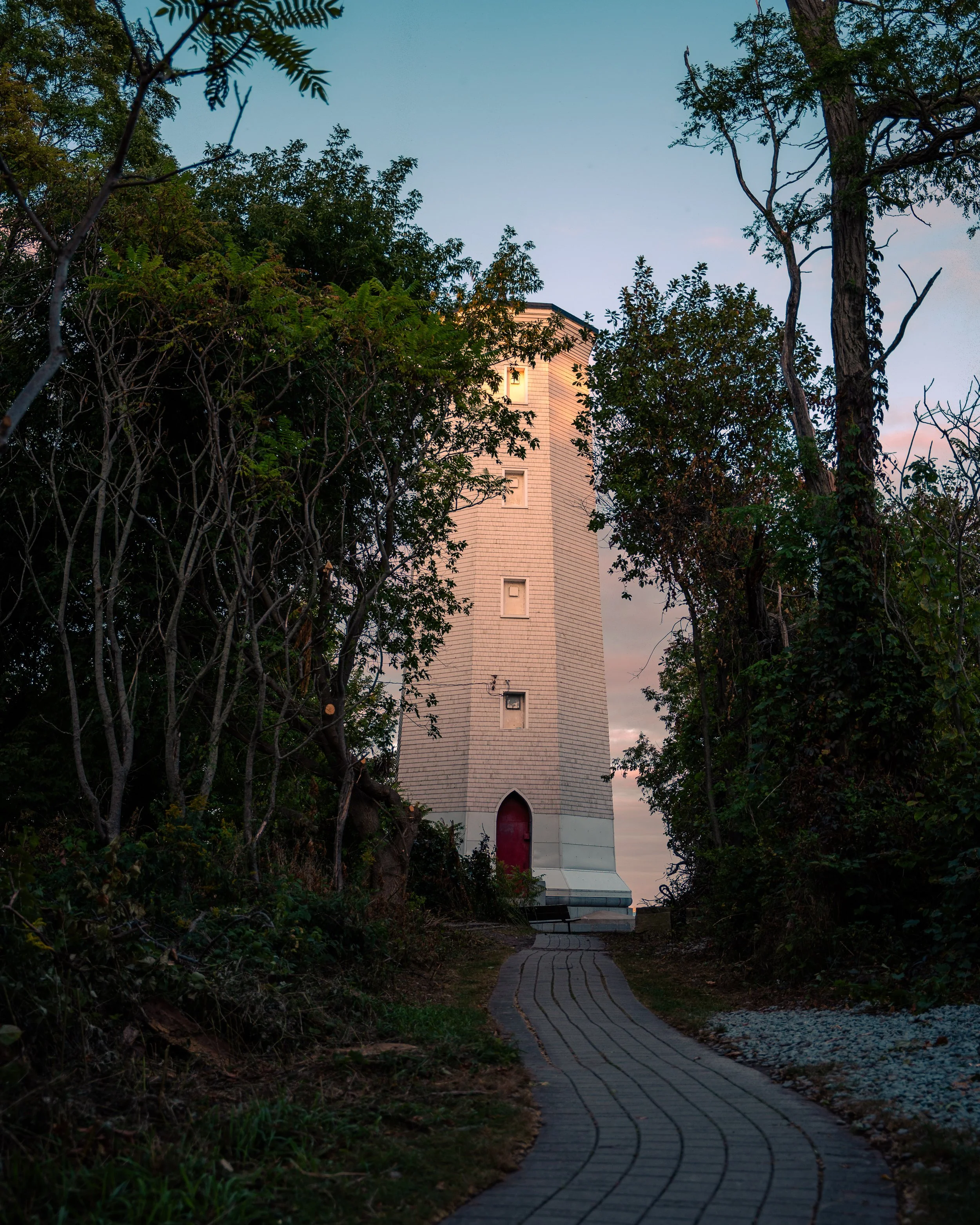 A winding paved pathway leads to a lighthouse or tower partially hidden by dense trees and vegetation at dusk or dawn.