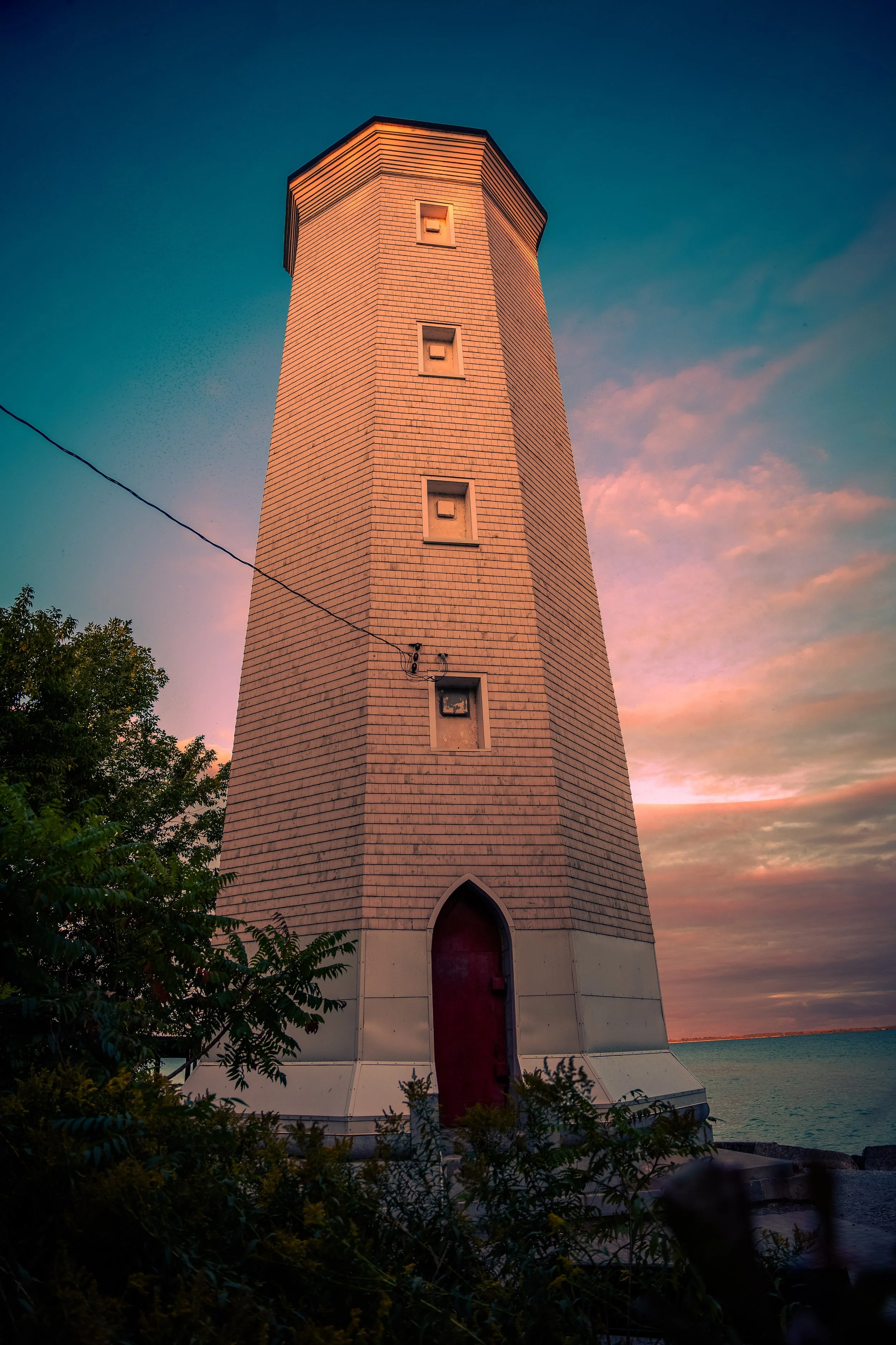 A lighthouse with a brick exterior painted white near the water at sunset, surrounded by trees and bushes.