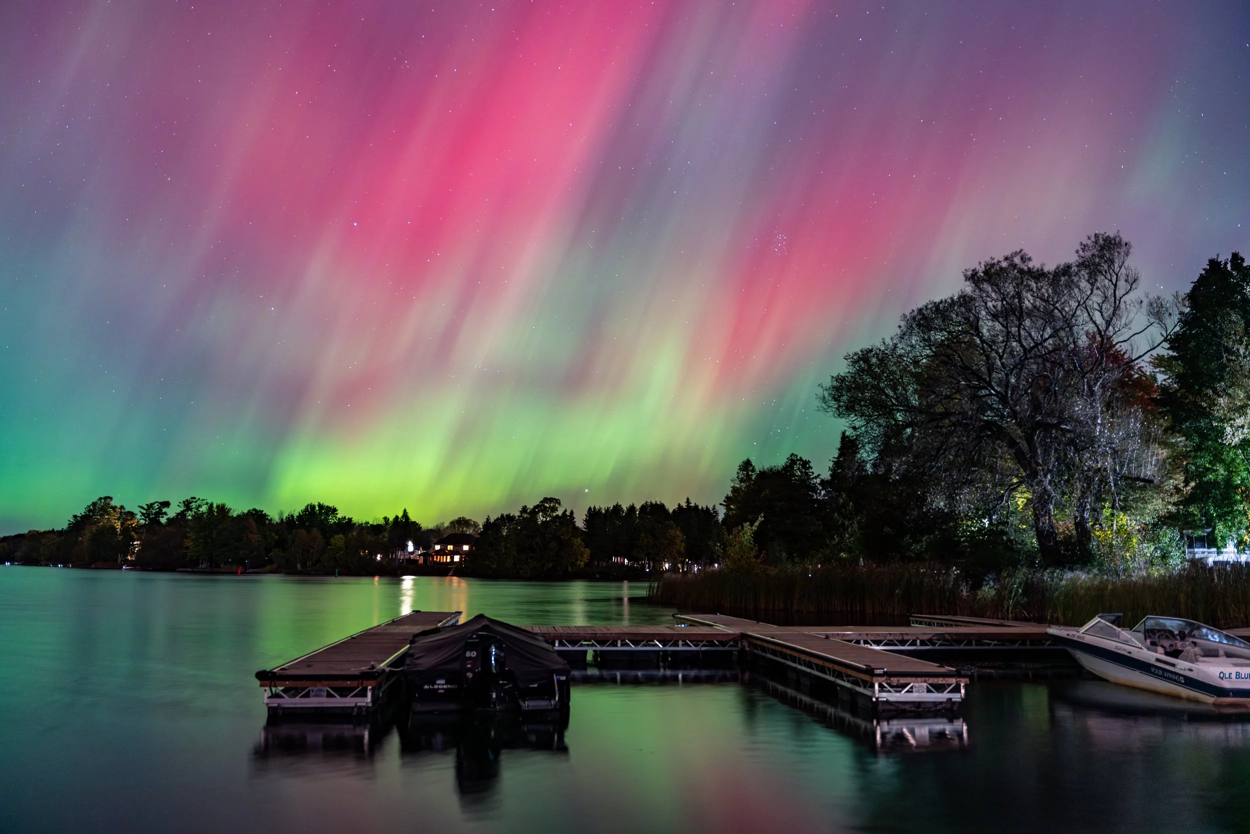 Northern lights illuminating the night sky over a lake, with trees and a dock in the foreground, and boats moored at the dock.