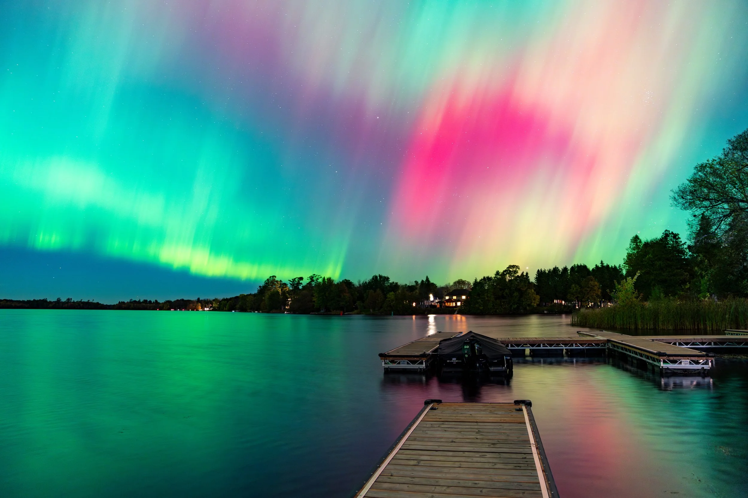 Colorful northern lights over a lake with a wooden dock and trees in the background.