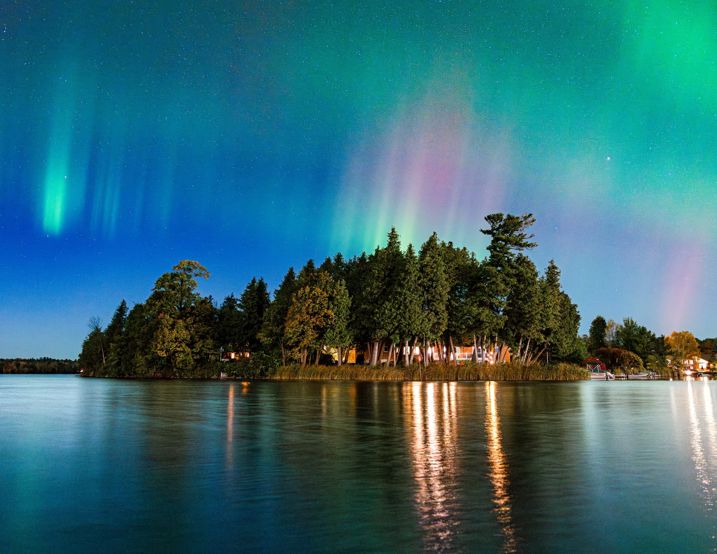 Night sky over a lake with the Northern Lights illuminating green and purple, with trees and a small house on the shoreline.