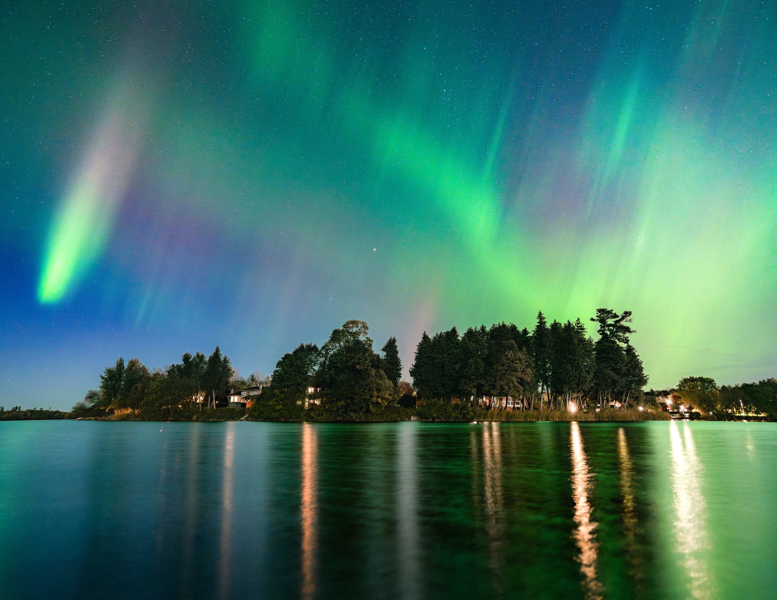 Northern Lights over a lake with trees and houses at night.