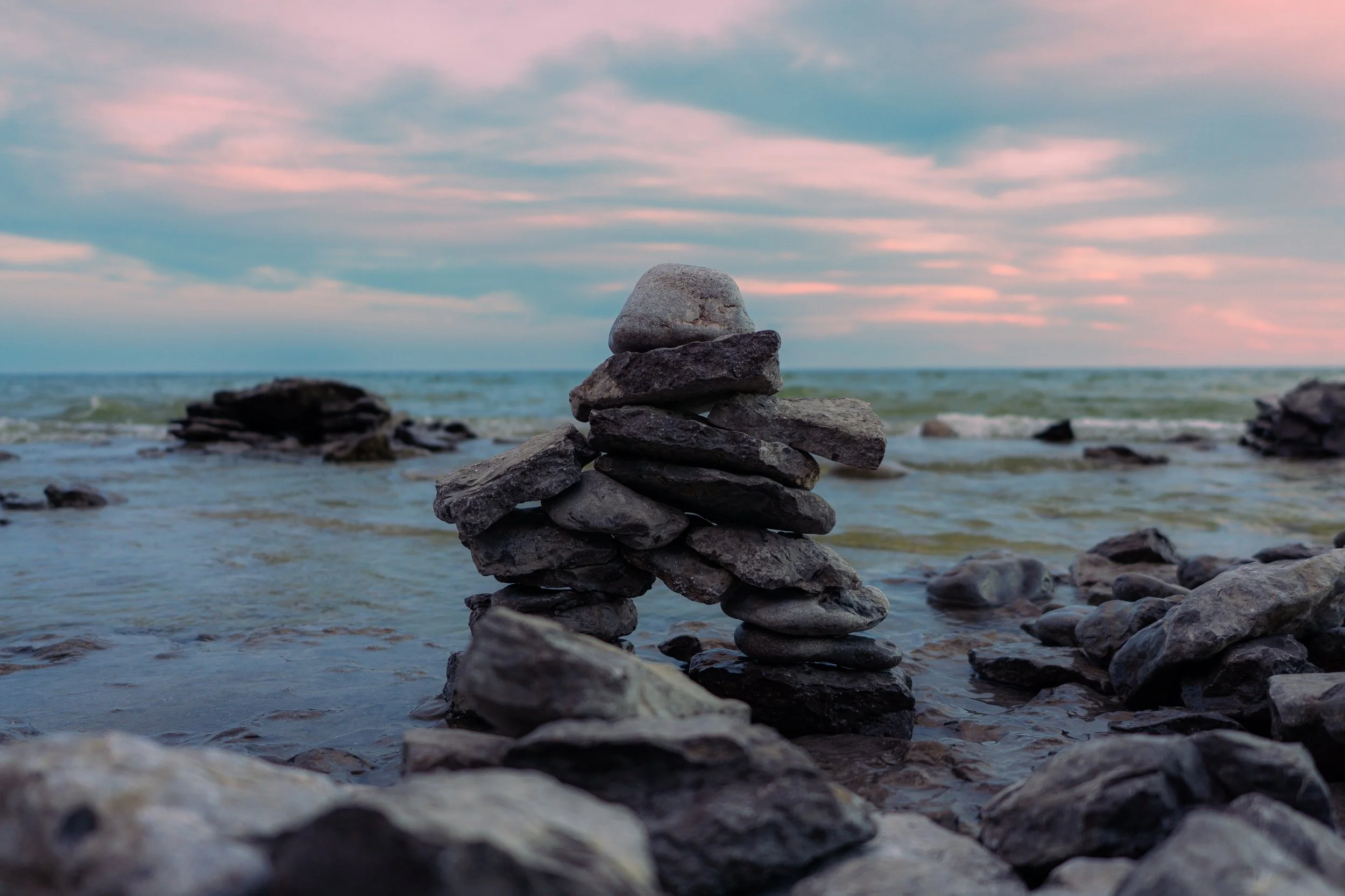 A stack of flat rocks arranged in a tower on a rocky beach with the ocean in the background and a sky with pink and blue clouds.