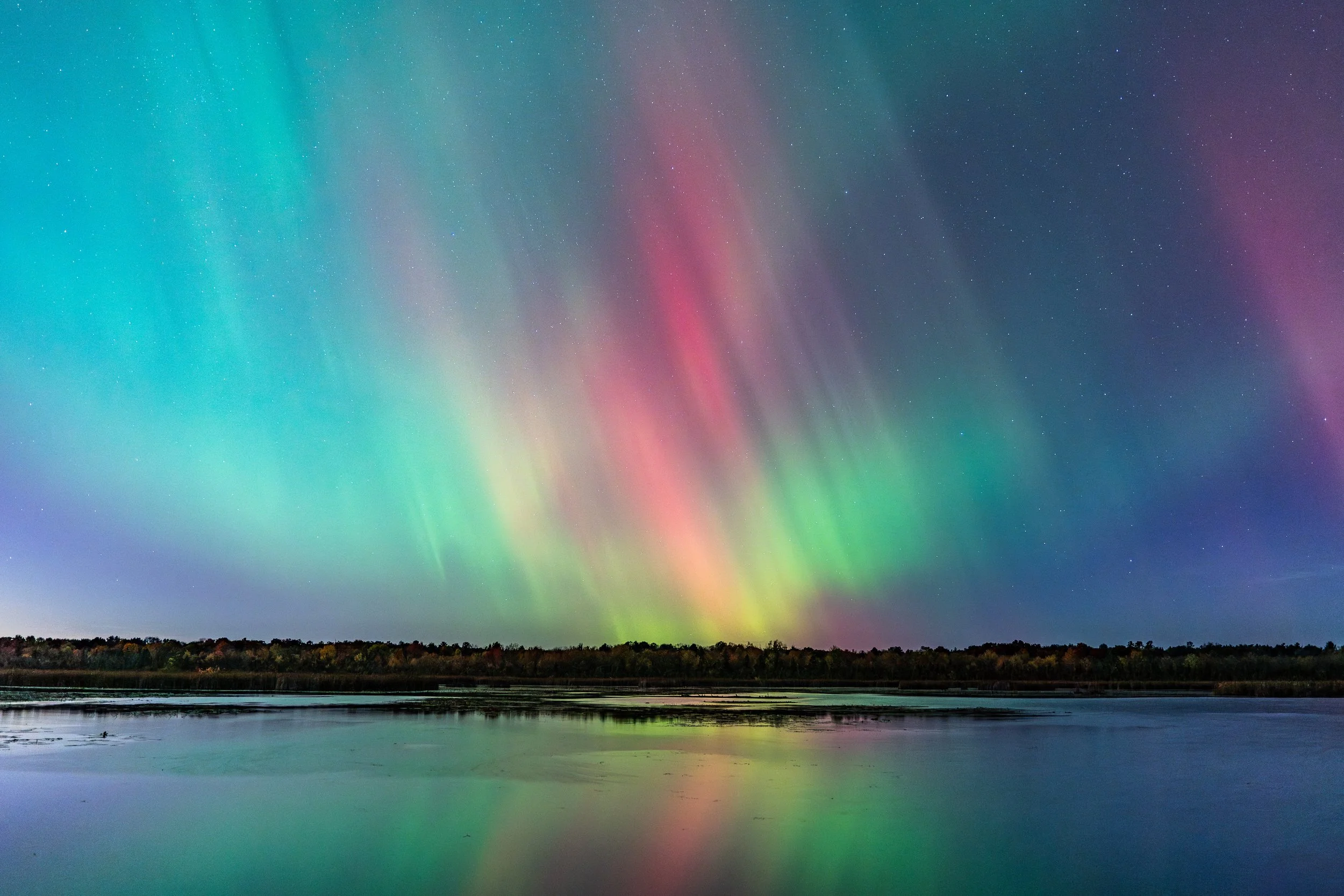 Northern lights illuminating the night sky over a calm lake with a forested shoreline.