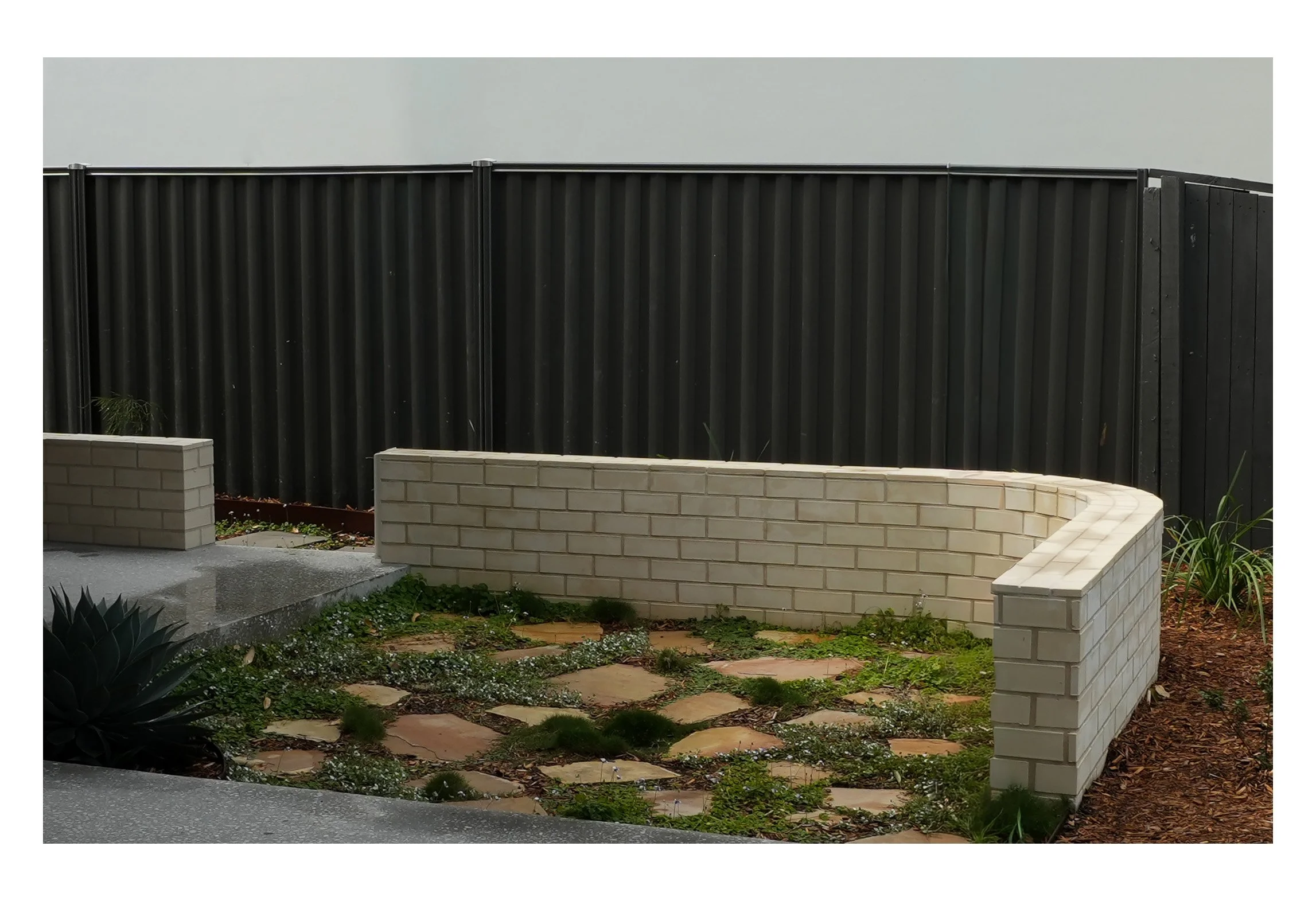 A backyard garden with a brick wall, a curved brick planter, and a black wooden fence in the background. There are some plants, including succulents and small shrubs, growing in the garden and along the walkway.