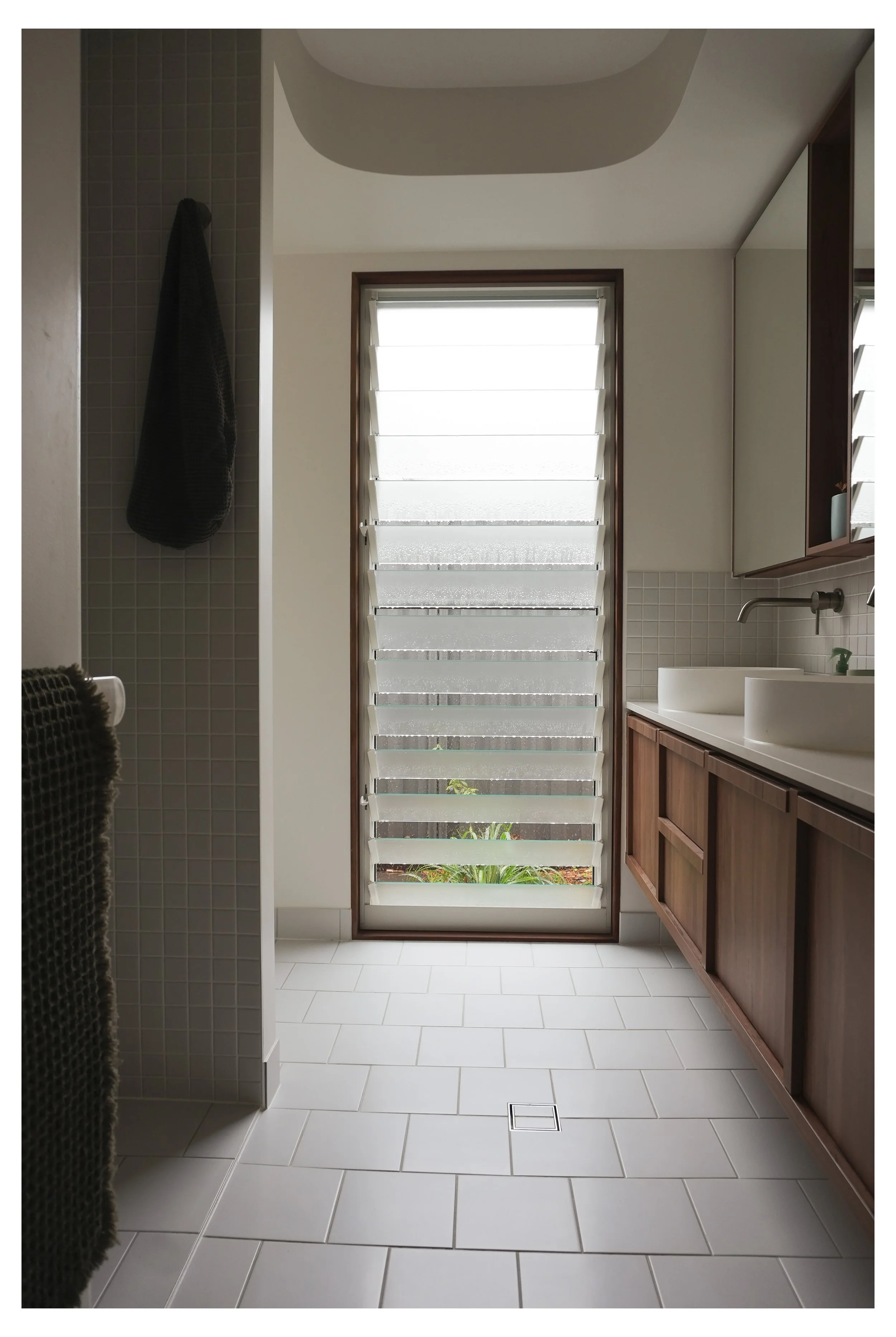 Modern bathroom with white rectangular tiles on the floor and beige small square tiles on the wall. A wooden vanity with two vessel sinks and a mirror cabinet. A window with adjustable white slats letting in natural light. Black hanging towel and a towel on the left. Indoor plants visible through the window.