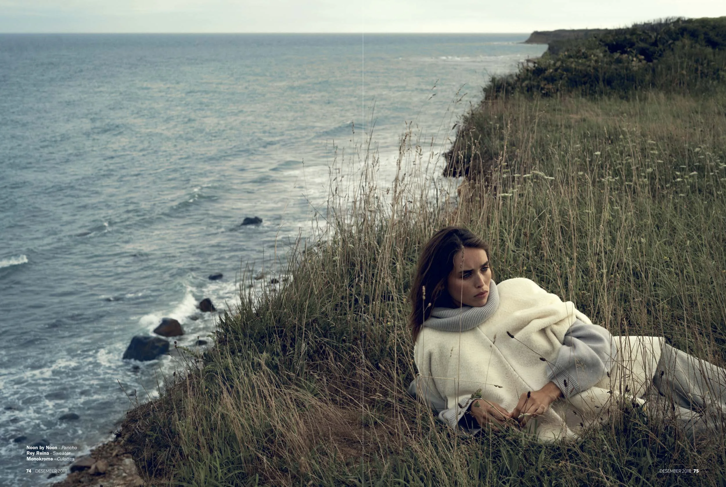 A woman with long brown hair, wearing a cream-colored sweater with a gray turtleneck collar, lies on a grassy cliff overlooking the ocean, with waves crashing on rocks below. She appears contemplative, gazing into the distance.