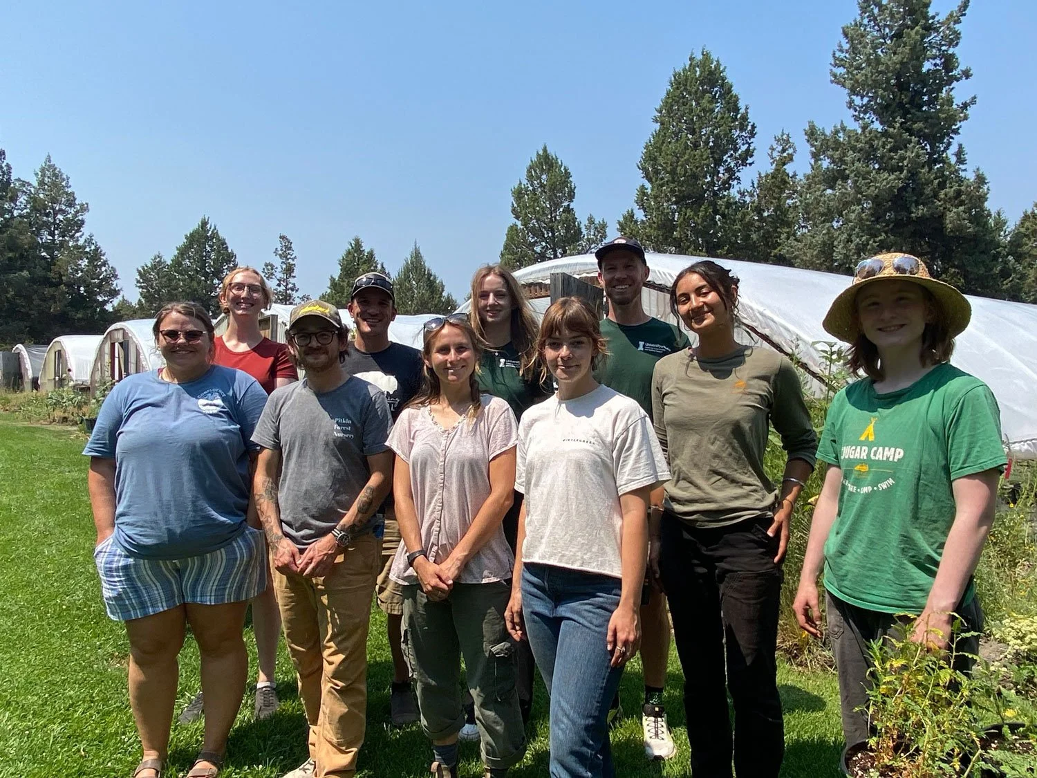 Group of people standing outdoors in front of greenhouses, smiling, on a sunny day with trees in the background.