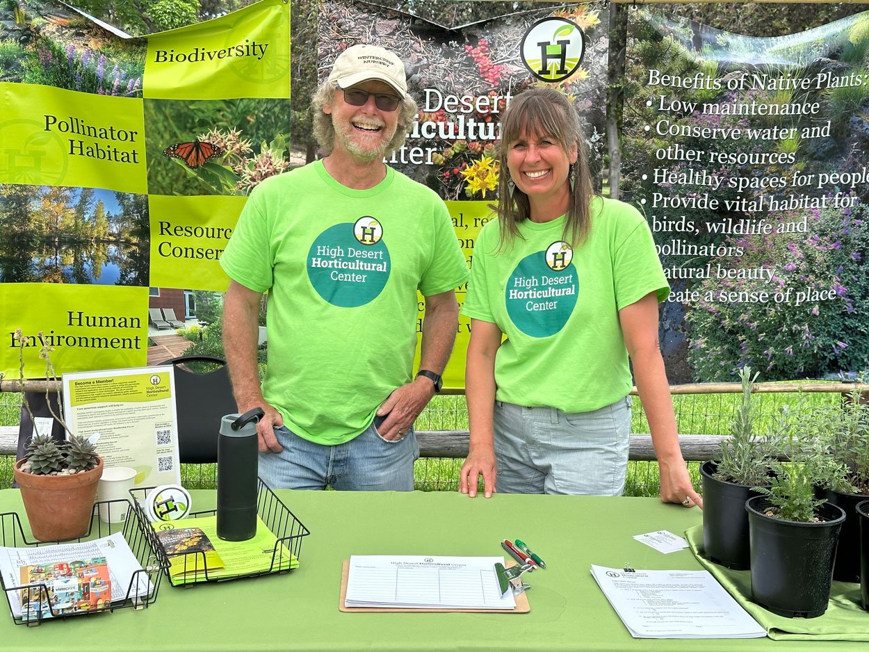 Two smiling people, a man and a woman, standing behind a table at a booth for the High Desert Horticultural Center. They are wearing bright green shirts with the organization's logo. The booth displays plants, informational materials, and a large banner promoting native plants and conservation.