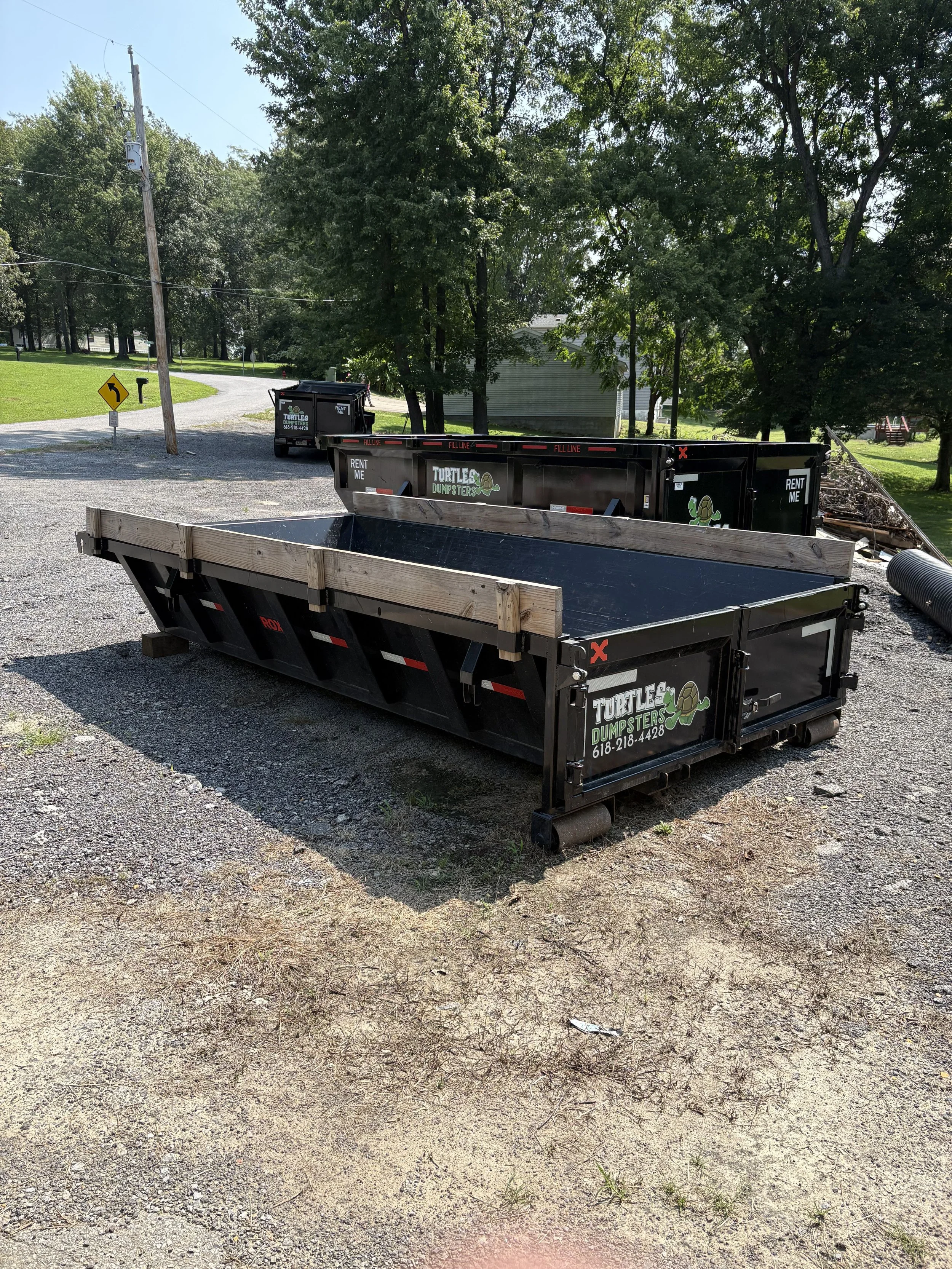 9 yard dumpster 'Turtles Dumpsters' with contact number, one filled with branches and debris, parked on a gravel driveway surrounded by green trees and grass.