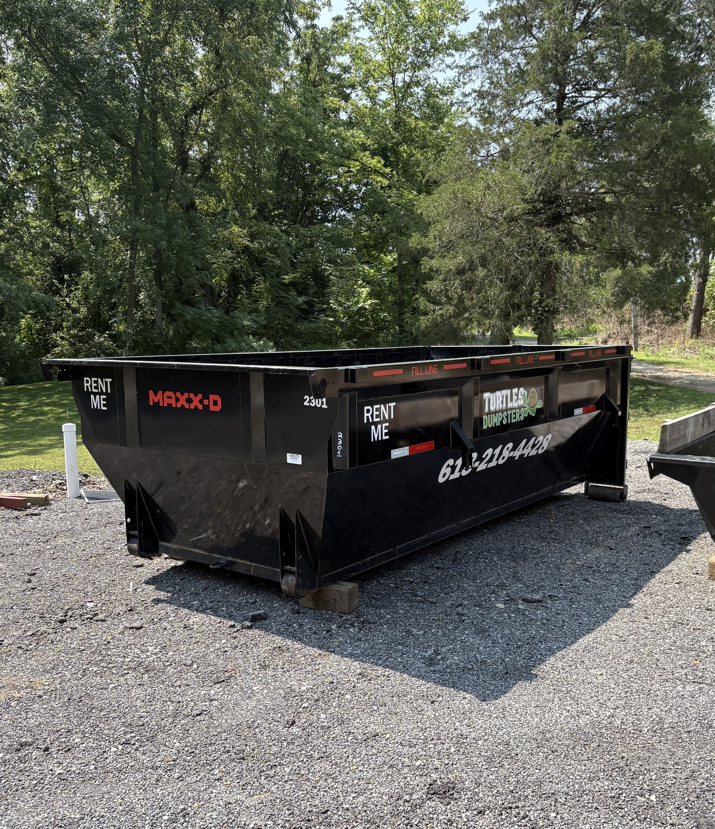 Empty 15 yard black dump trailer with 'Turtle Dumpsters' branding, parked on a gravel surface with trees in the background.